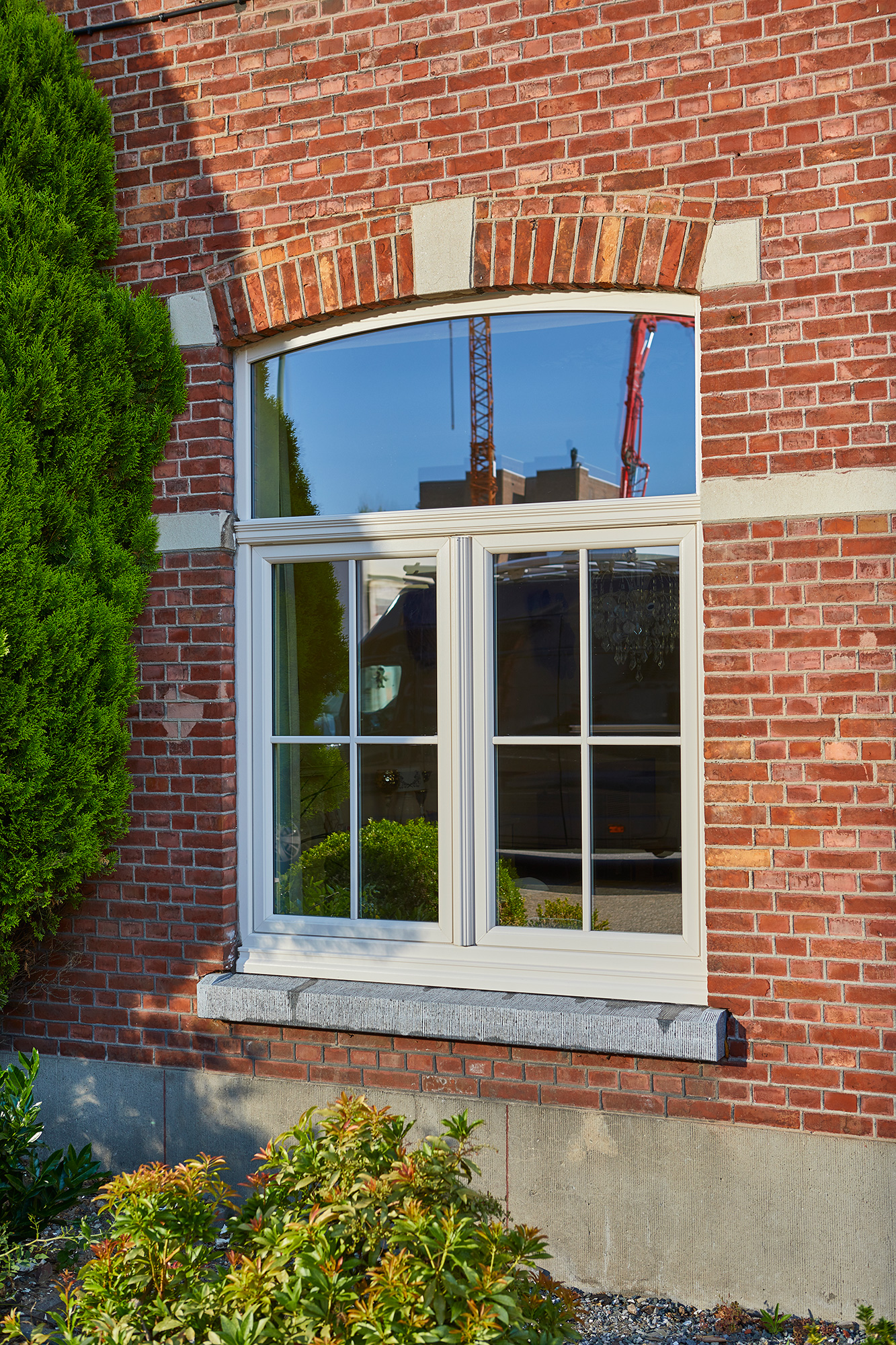 Crèmekleurig raam in verfijnd klassieke stijl, verwerkt in de bakstenen gevel van een gerenoveerde gesloten bebouwing, omgeven door groen.