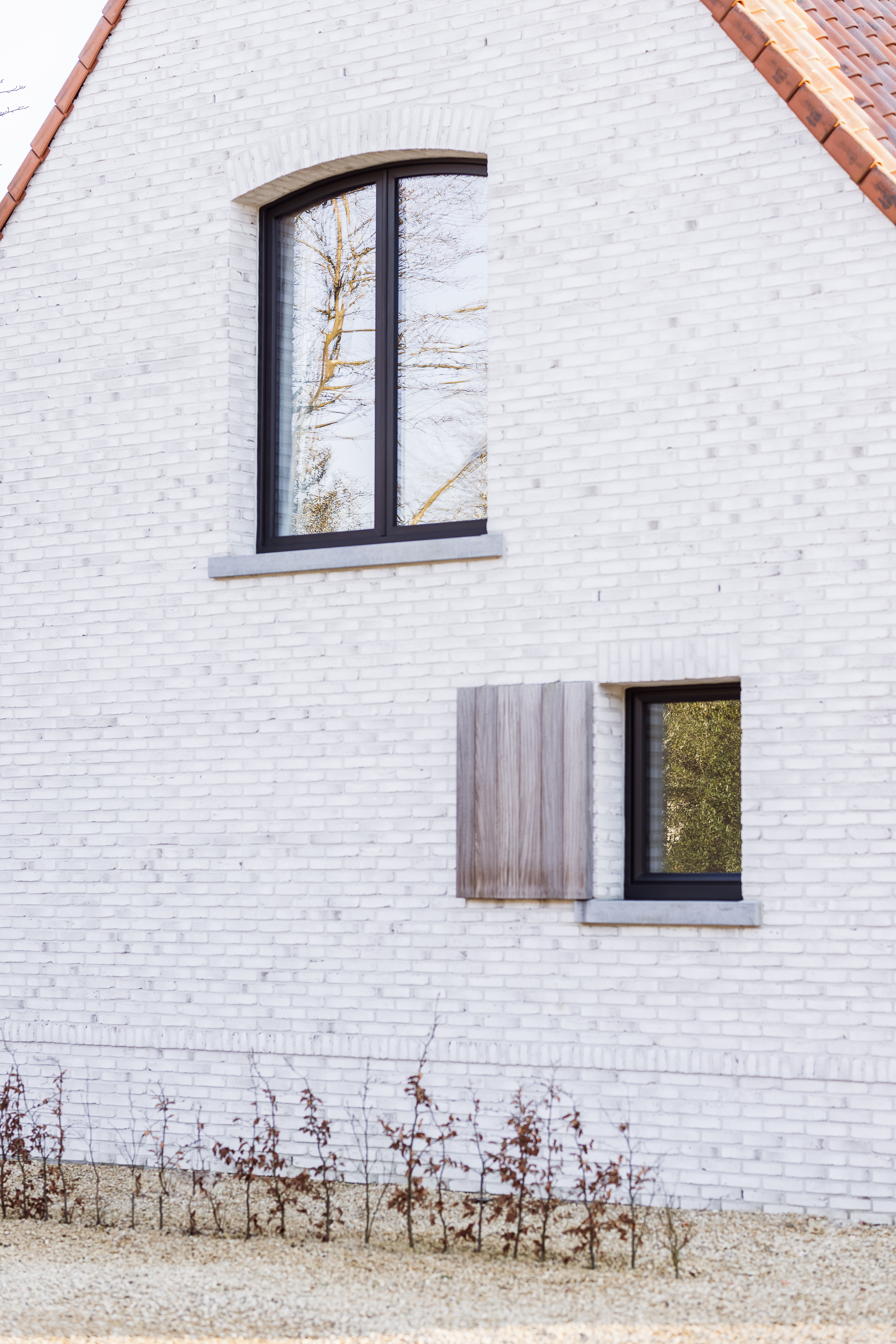 Een lichte bakstenen gevel met een hoog, rondboogvormig Impakt PLUS rond-raam in mat zwart en een kleiner raam met houten luik eronder.