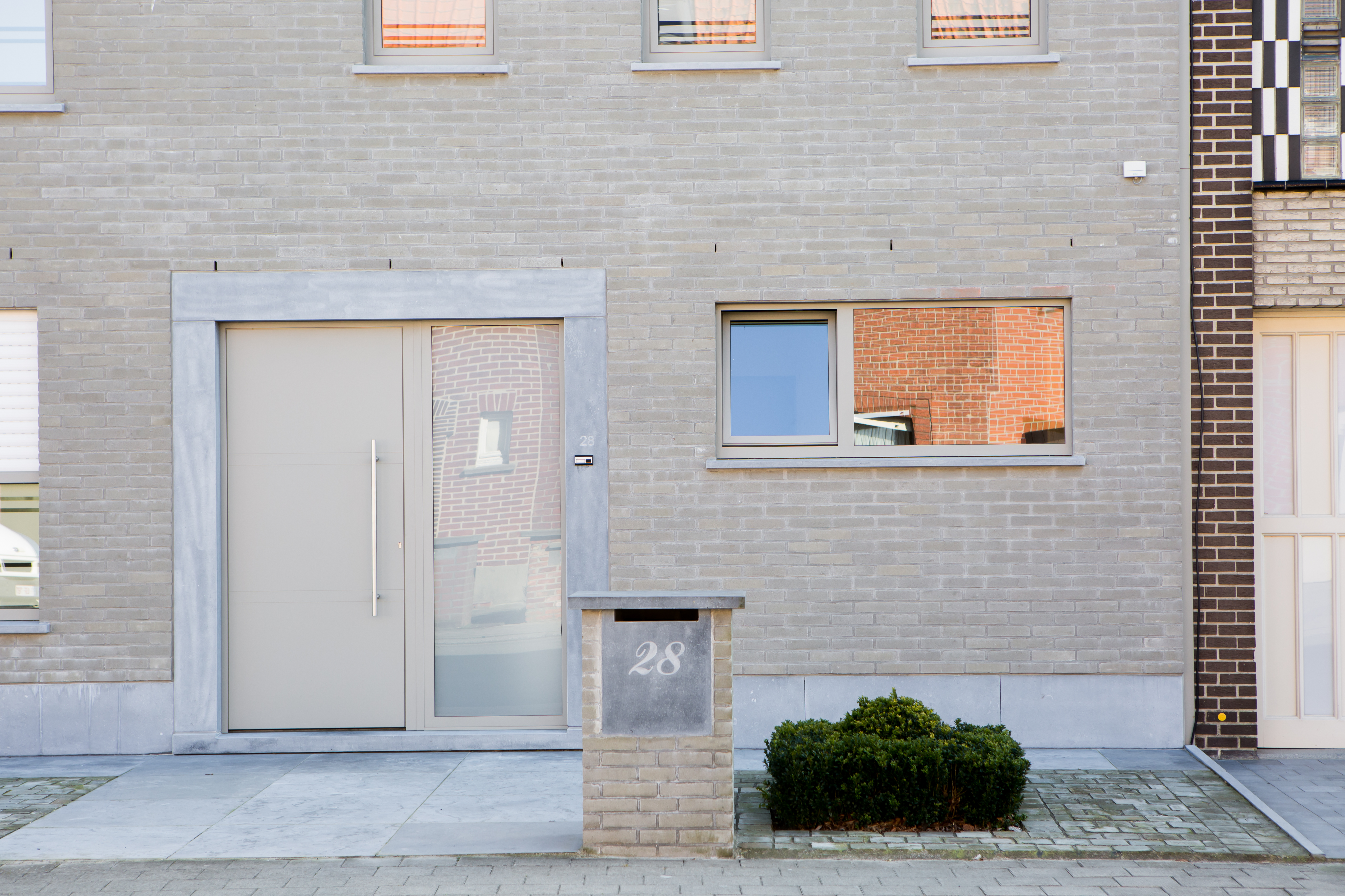 Grijze bakstenen gevel van een driegevelwoning met een semi-beglaasde voordeur, brievenbus met huisnummer 28 en een kleine voortuin.