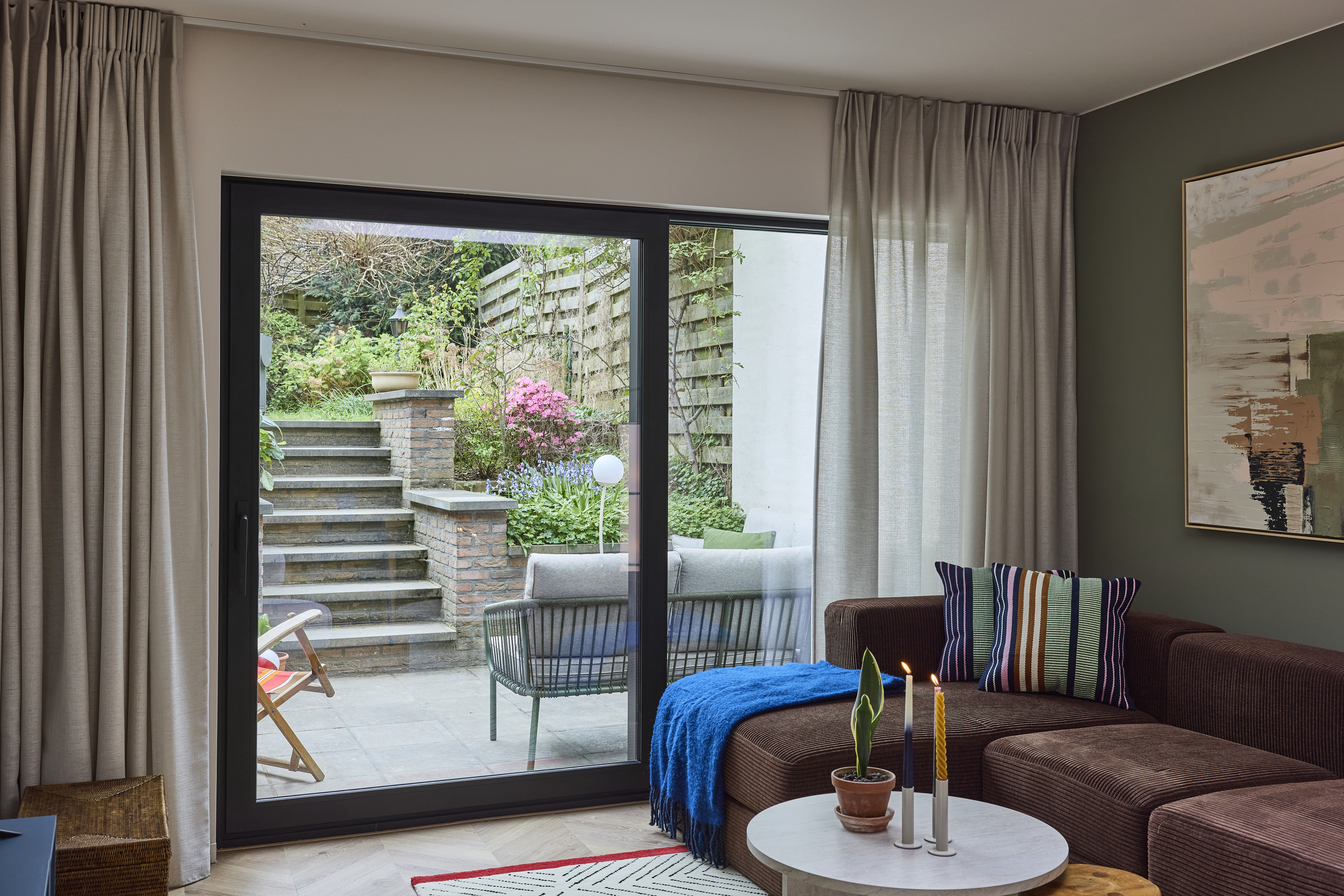 Zwart ALU Visoglide Monorail-schuifraam in een woonkamer, met glas tot op de vloer en zicht op een terras met trap en tuin.