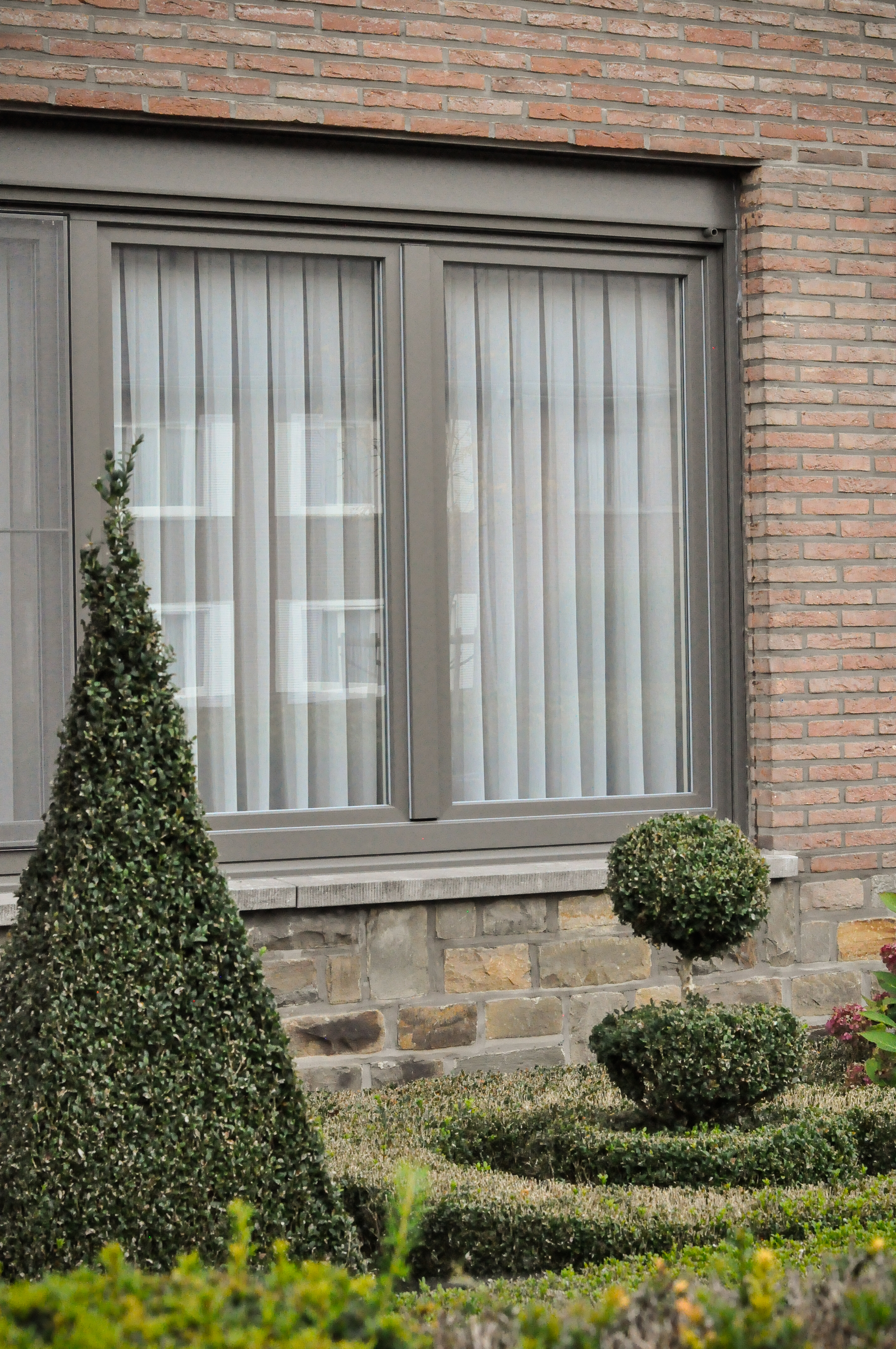 Gerenoveerde, tijdloze halfopen woning met grijze raamprofielen en een groot raam met drie verticale delen in een bakstenen gevel, naast een strak gesnoeide tuin.