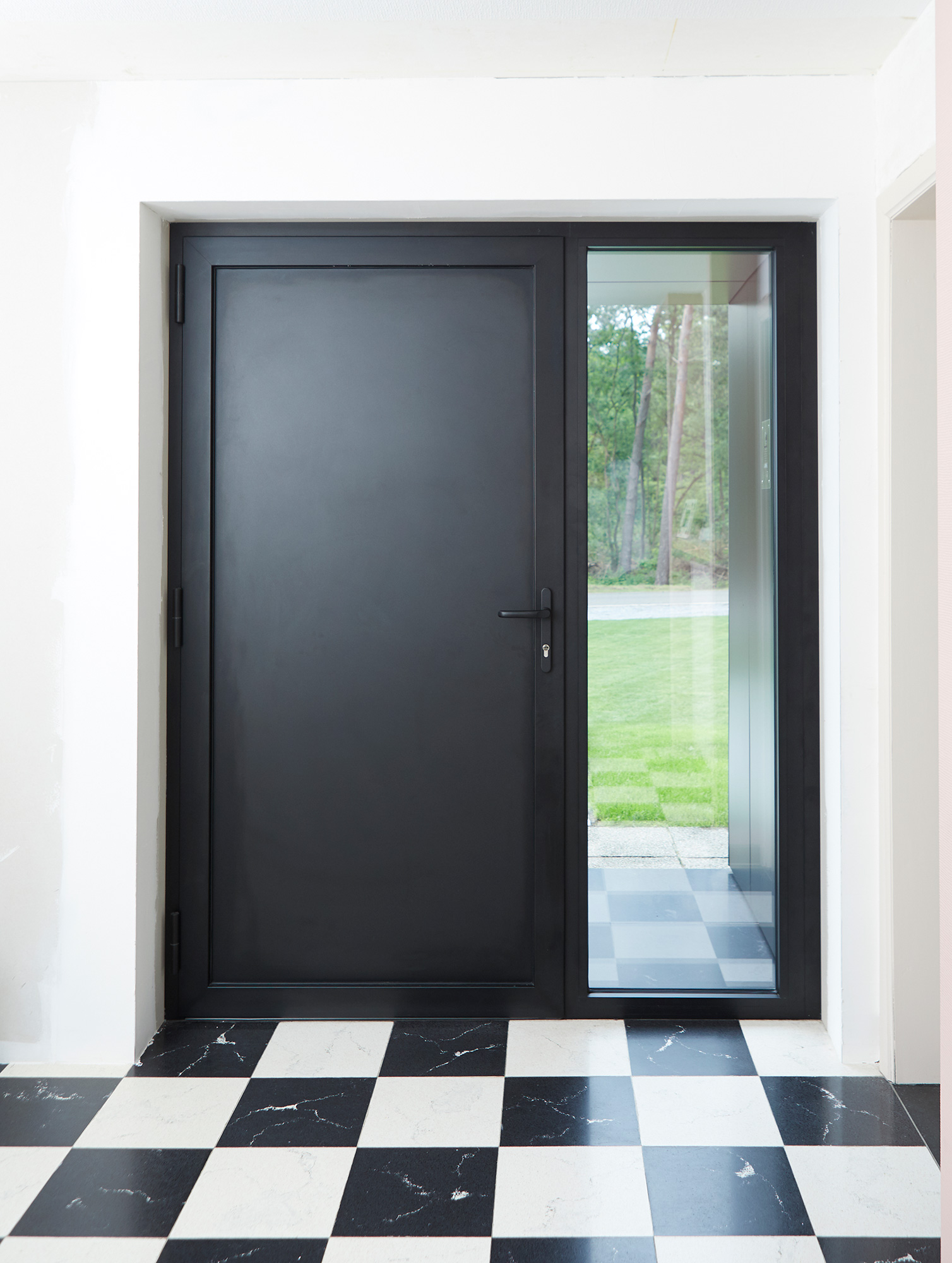 Moderne zwarte steellook-buitendeur met smal zijlicht in een gerenoveerde, lichte inkomhal met zwart-wit geblokte tegelvloer.