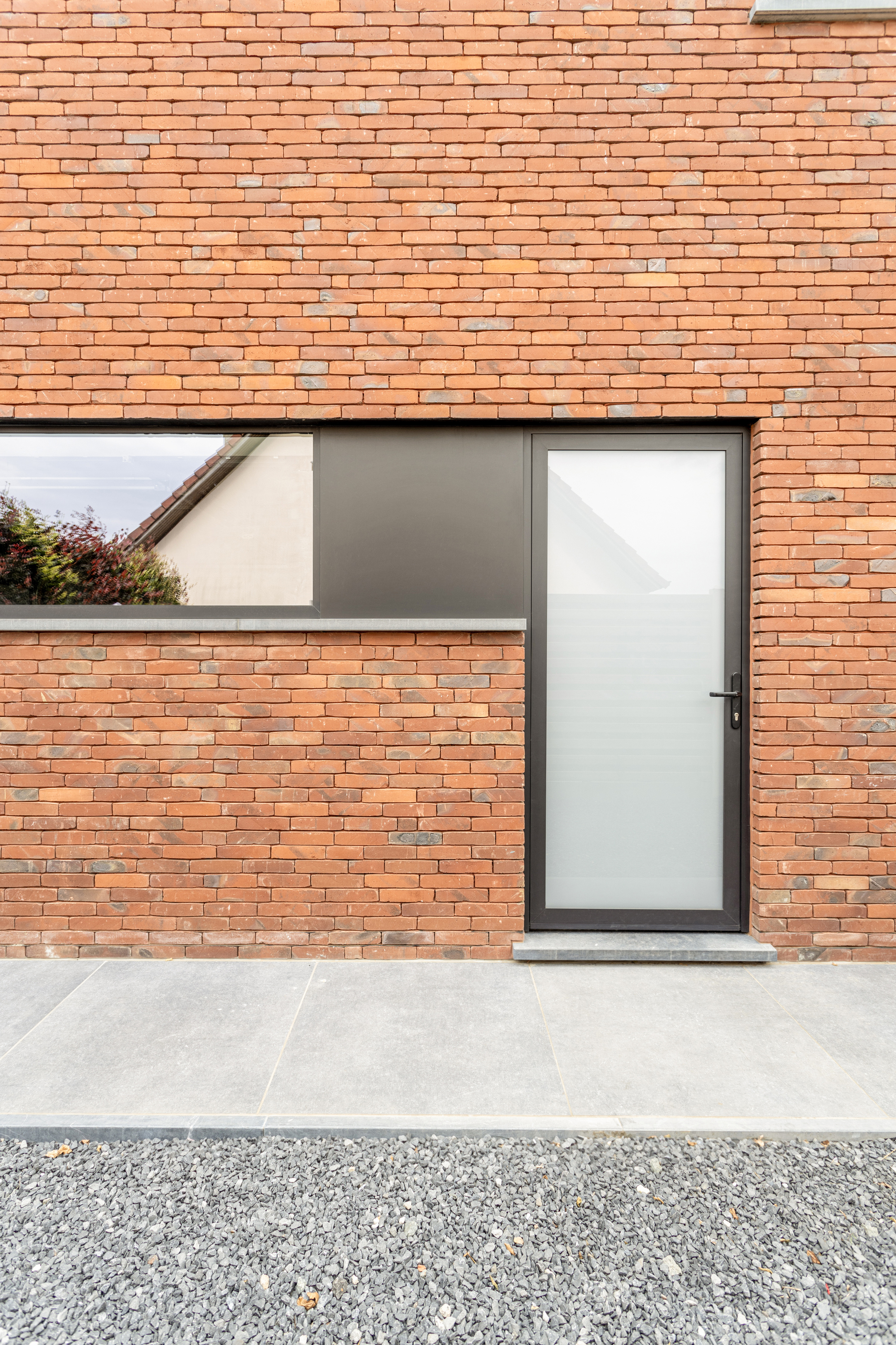 Zwarte, beglaasde aluminiumdeur met een moderne uitstraling in een rode bakstenen gevel van een nieuwbouwwoning, naast een smal raam.