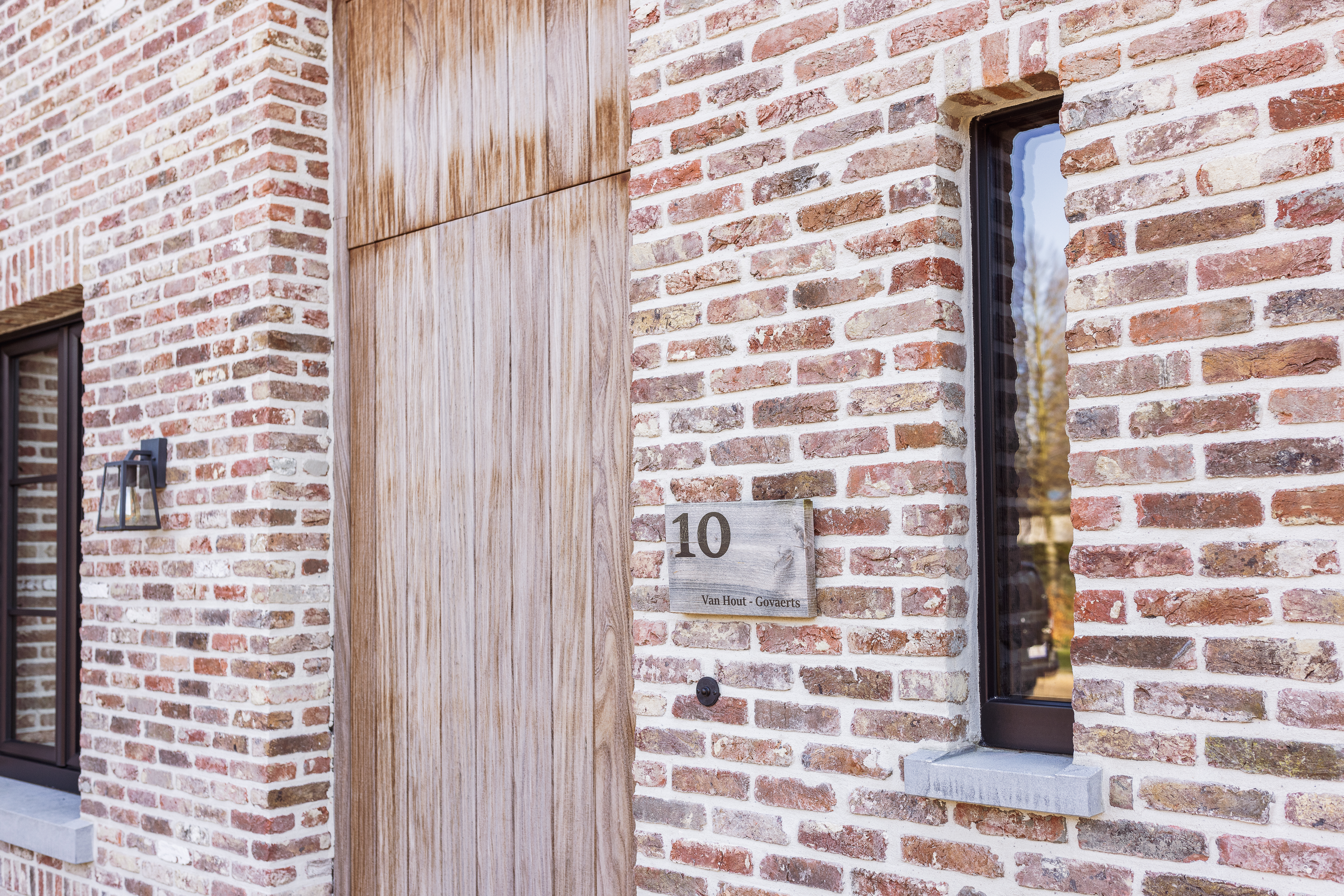 Bakstenen gevel met houten voordeur, zwart raamprofiel, huisnummer 10 en een zwarte ronde deurbel naast de inkom.