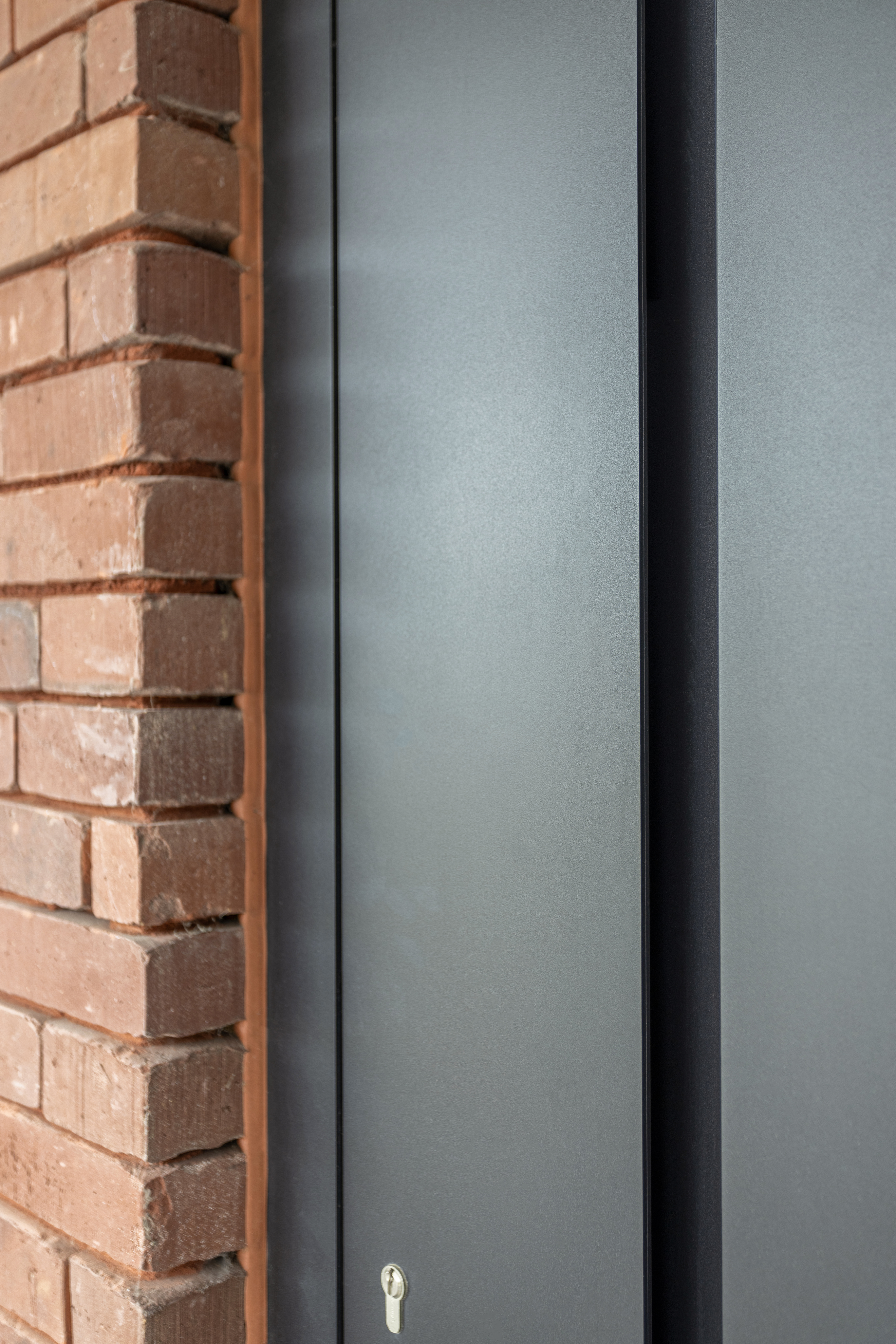 Modern zwart aluminiumpaneel naast een karaktervolle bakstenen gevel, met een smalle verticale naad en zichtbaar sleutelgat.