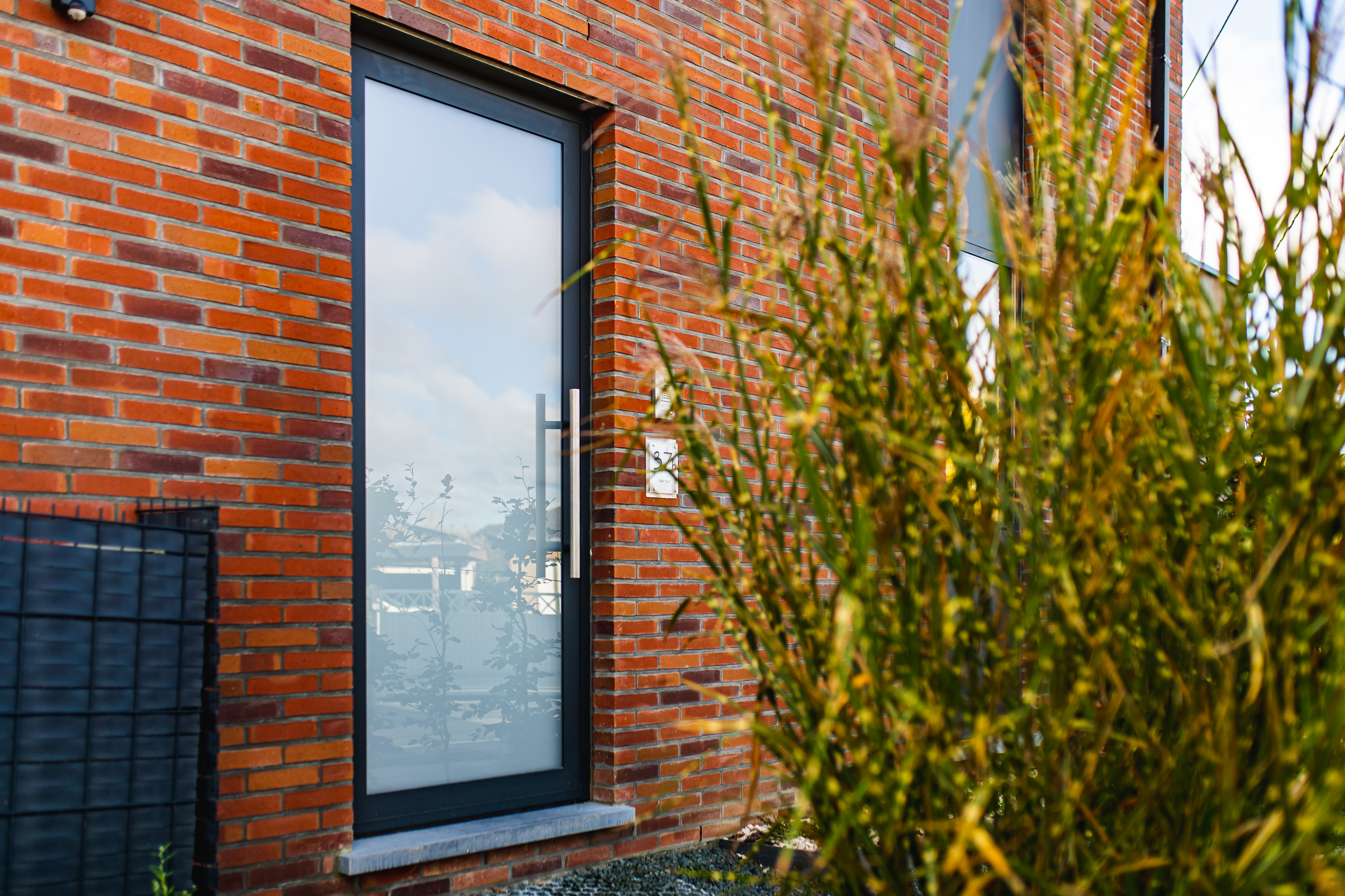 Smal antracietgrijs raam met donker raamprofiel in de rode bakstenen gevel van een vrijstaande nieuwbouwwoning, gedeeltelijk verscholen achter siergrassen.