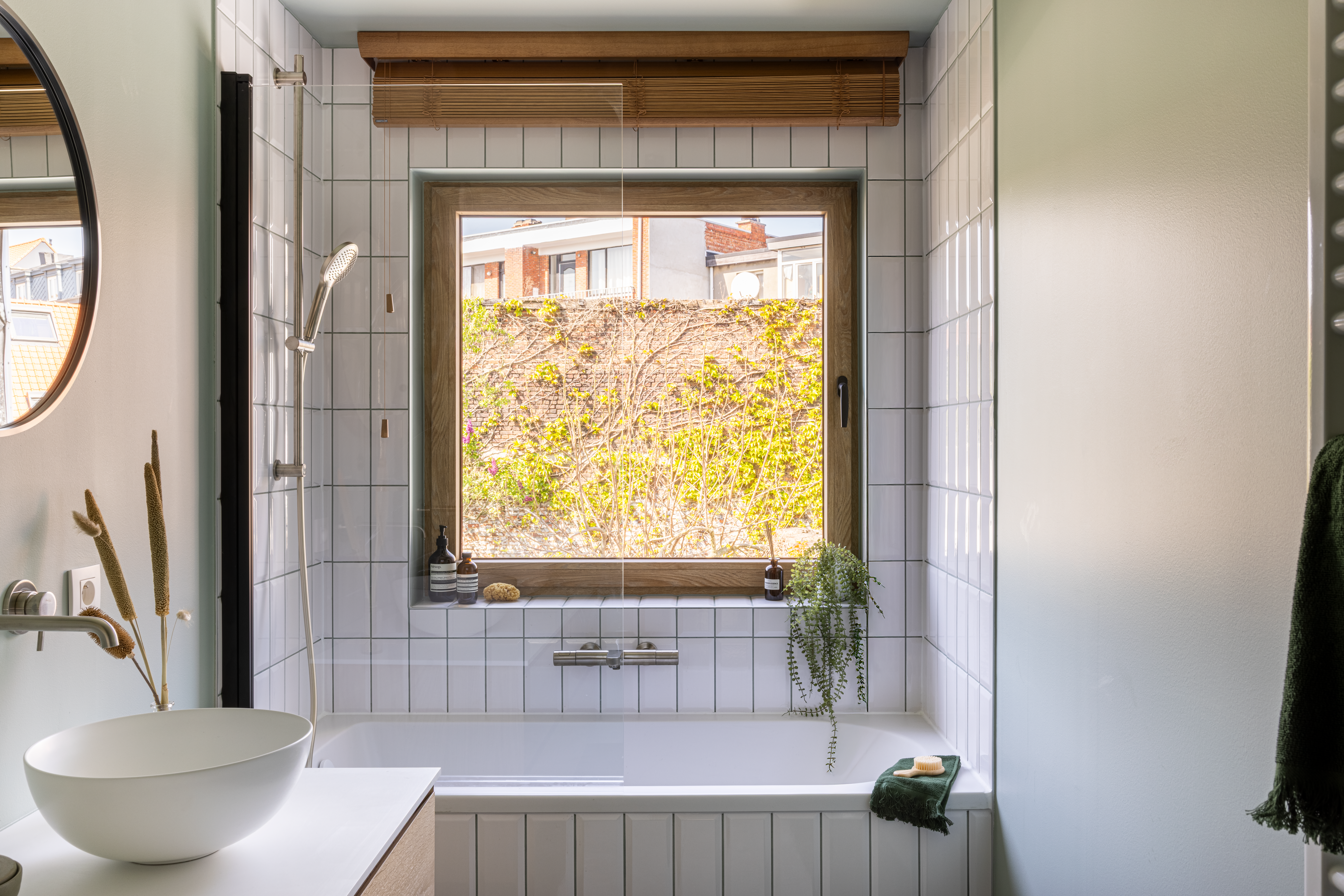 Draai-kipraam met natuurlijke eiklook boven een bad, in een lichte badkamer met witte wandtegels, houten jaloezie en groene plant.
