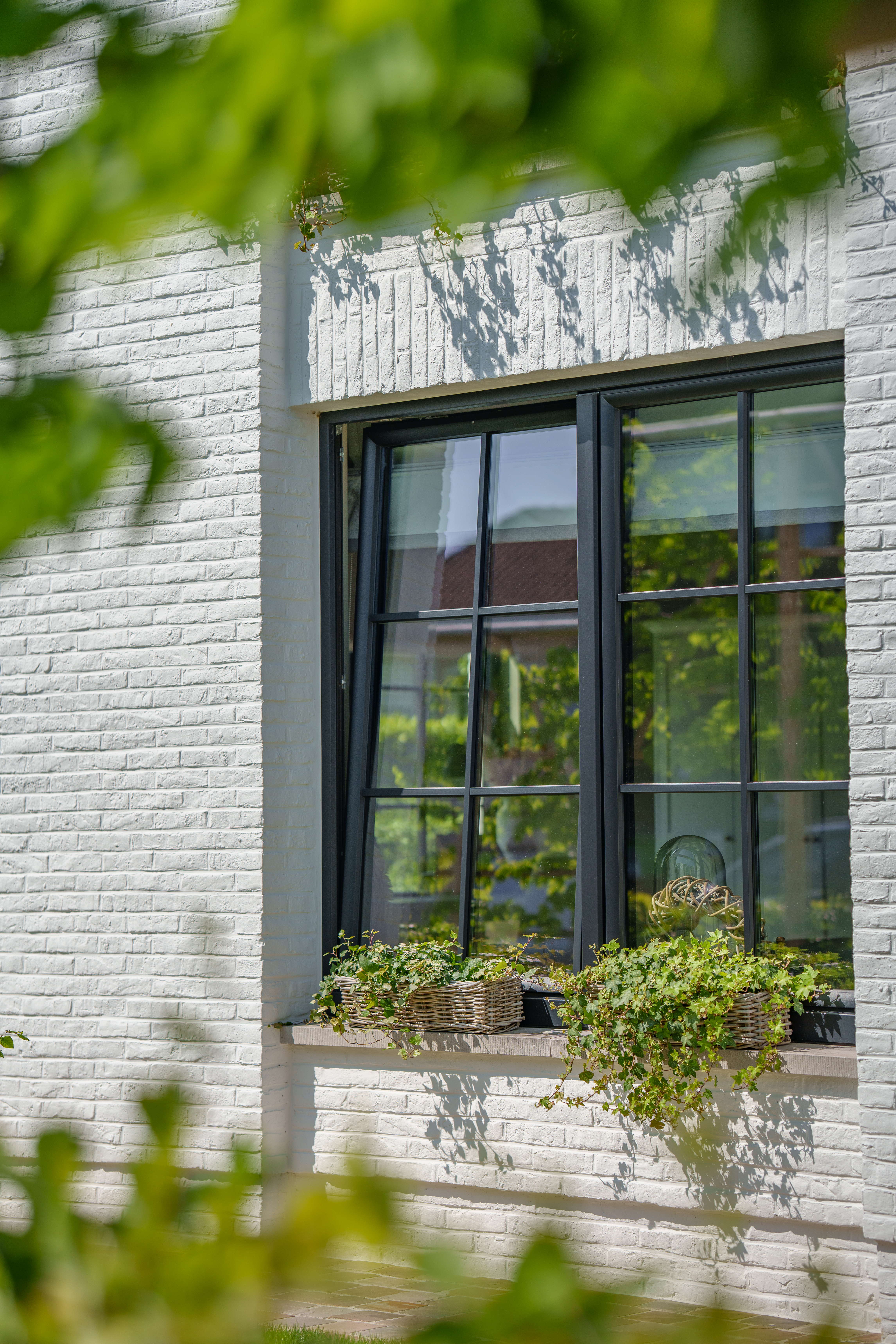 Zwart mat dubbel raam met draai-kipfunctie in een lichte bakstenen gevel, met donkere raamprofielen, roedeverdeling en een begroeide vensterbank in warme landelijke stijl.