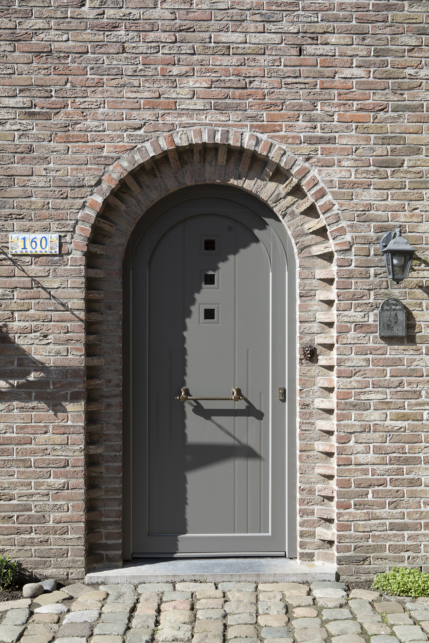 Grijze rondboogdeur uit de Impakt PLUS rond-reeks in een bakstenen gevel, met zwarte deurklopper, huisnummer 160 en buitenverlichting.