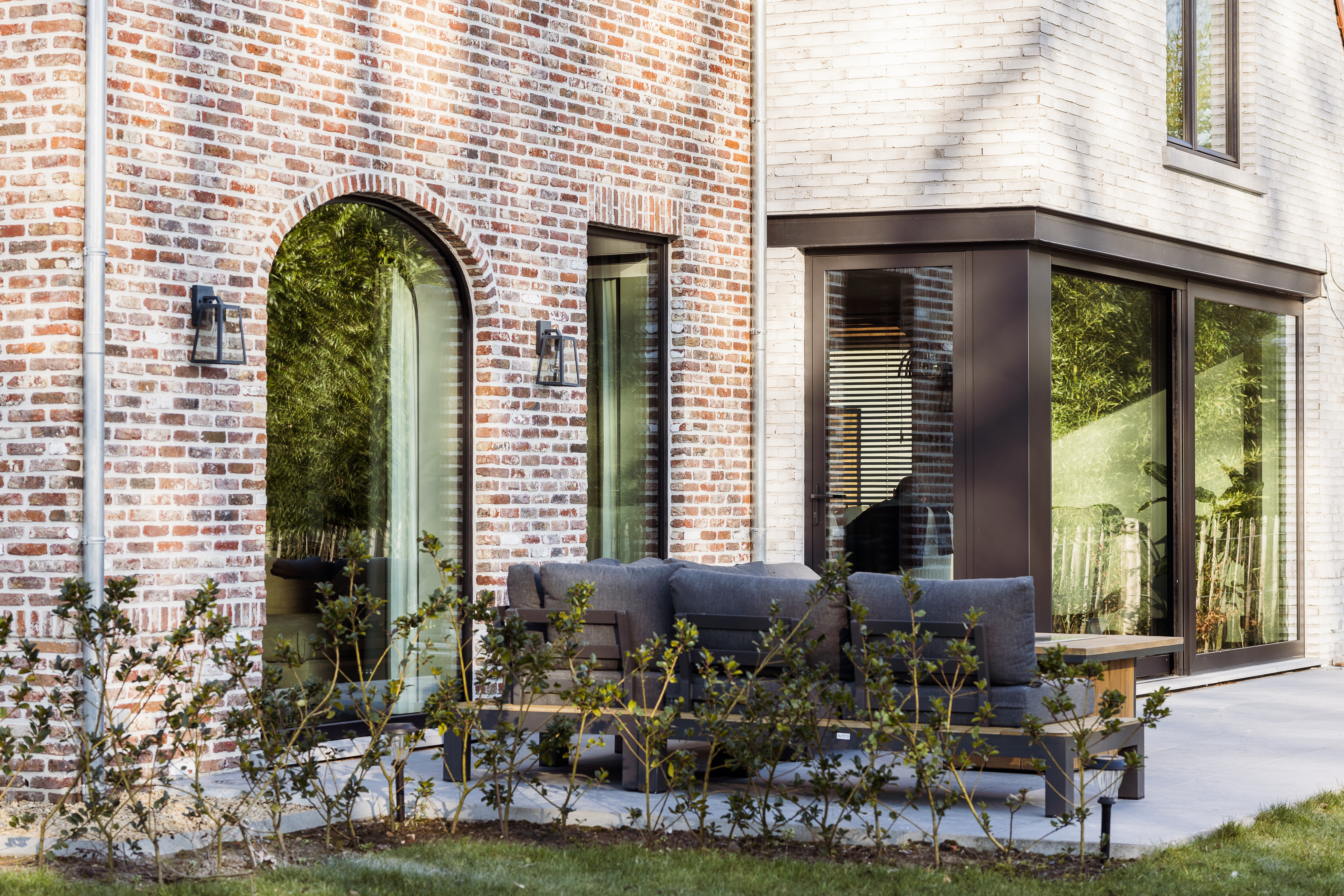 Moderne bakstenen gevel met matzwarte raamprofielen, een rondboograam en grote hoekbeglazing naast een terras met tuinmeubelen.