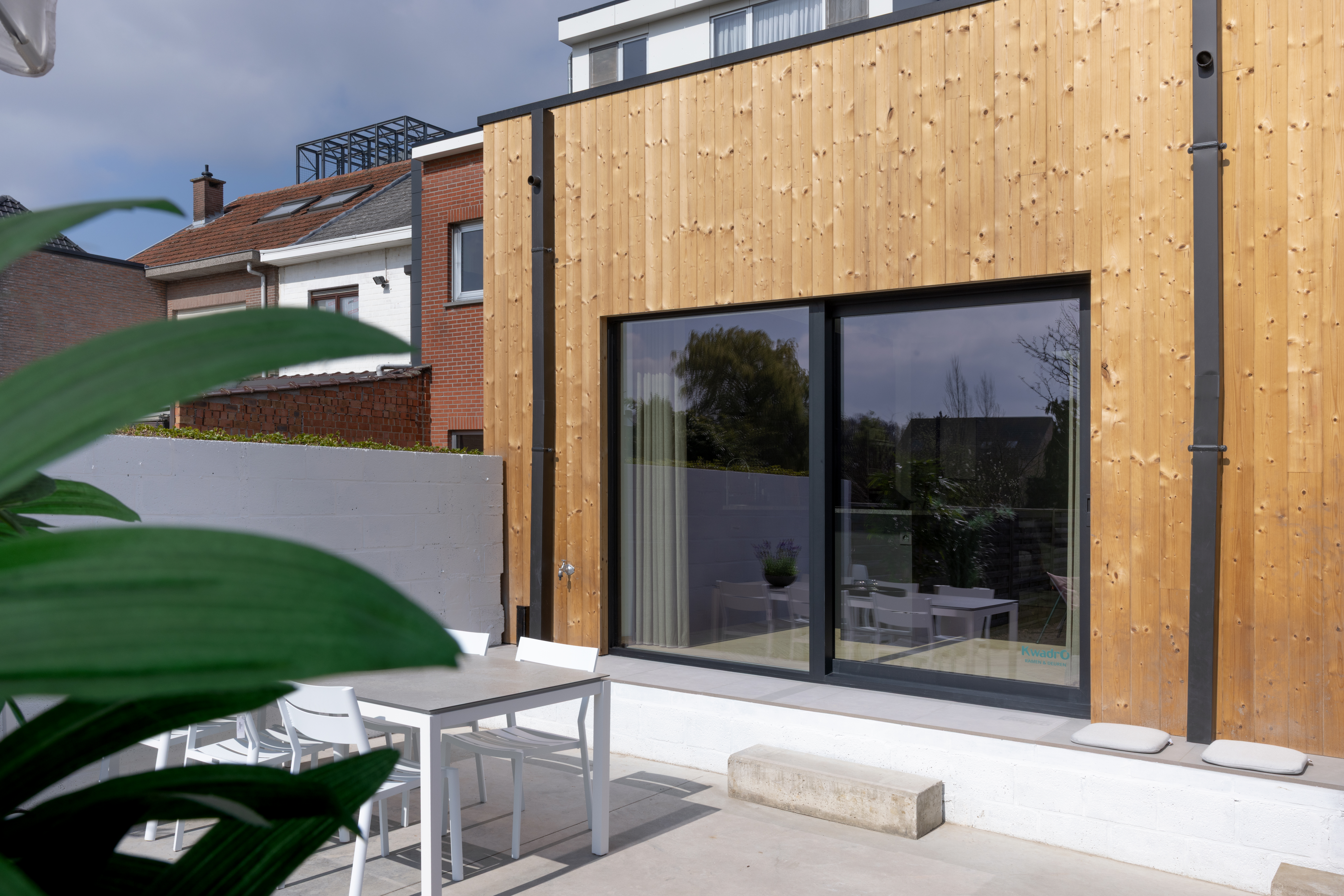 Moderne aanbouw met houten gevelbekleding en een groot grafietzwart aluminium schuifraam van het type ALU Visoglide Monorail, naast een terras.