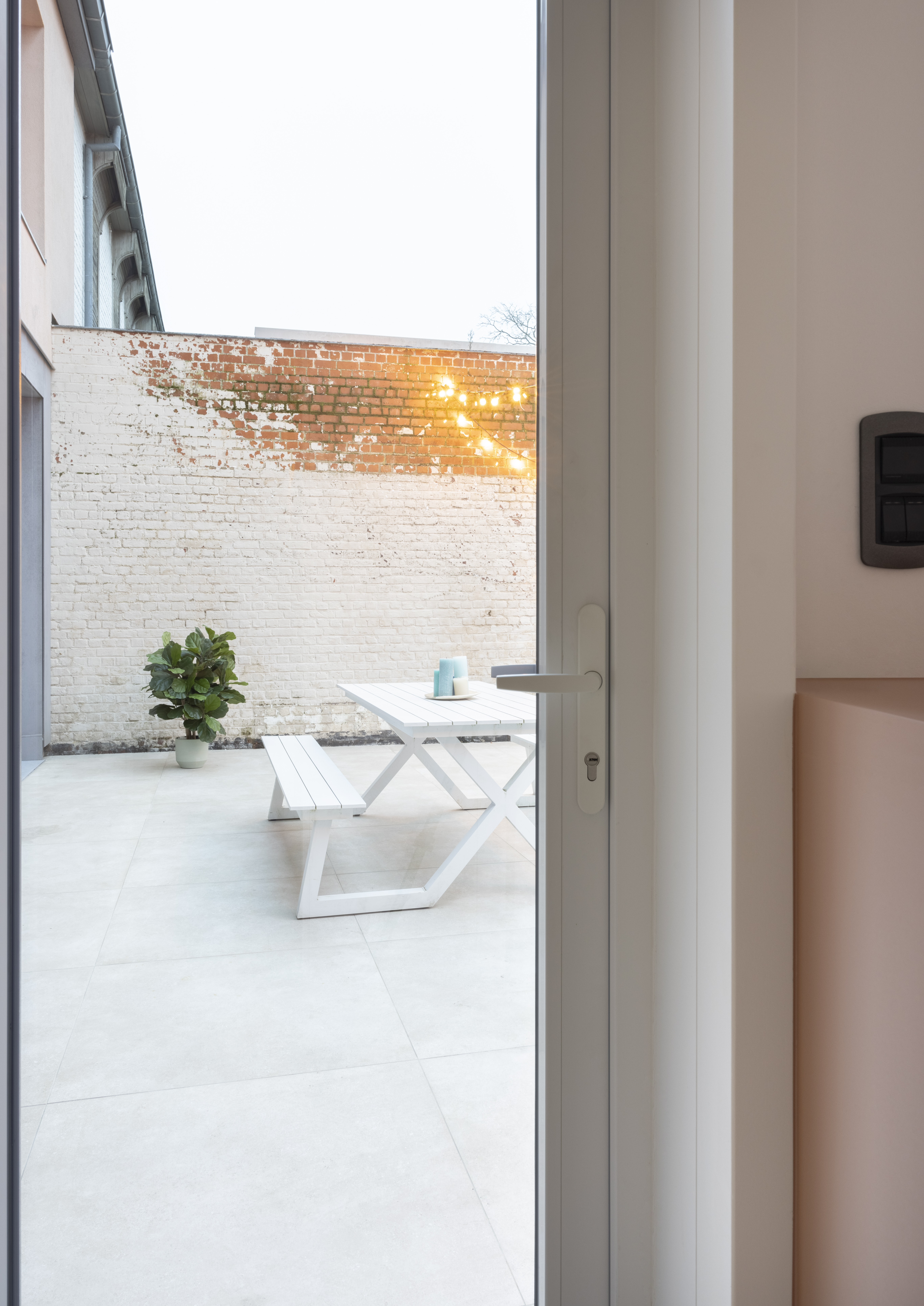Witte, moderne beglaasde pvc-deur met zicht vanuit het interieur op een lichte binnenkoer, een bakstenen muur, een plant en witte tuinmeubelen.