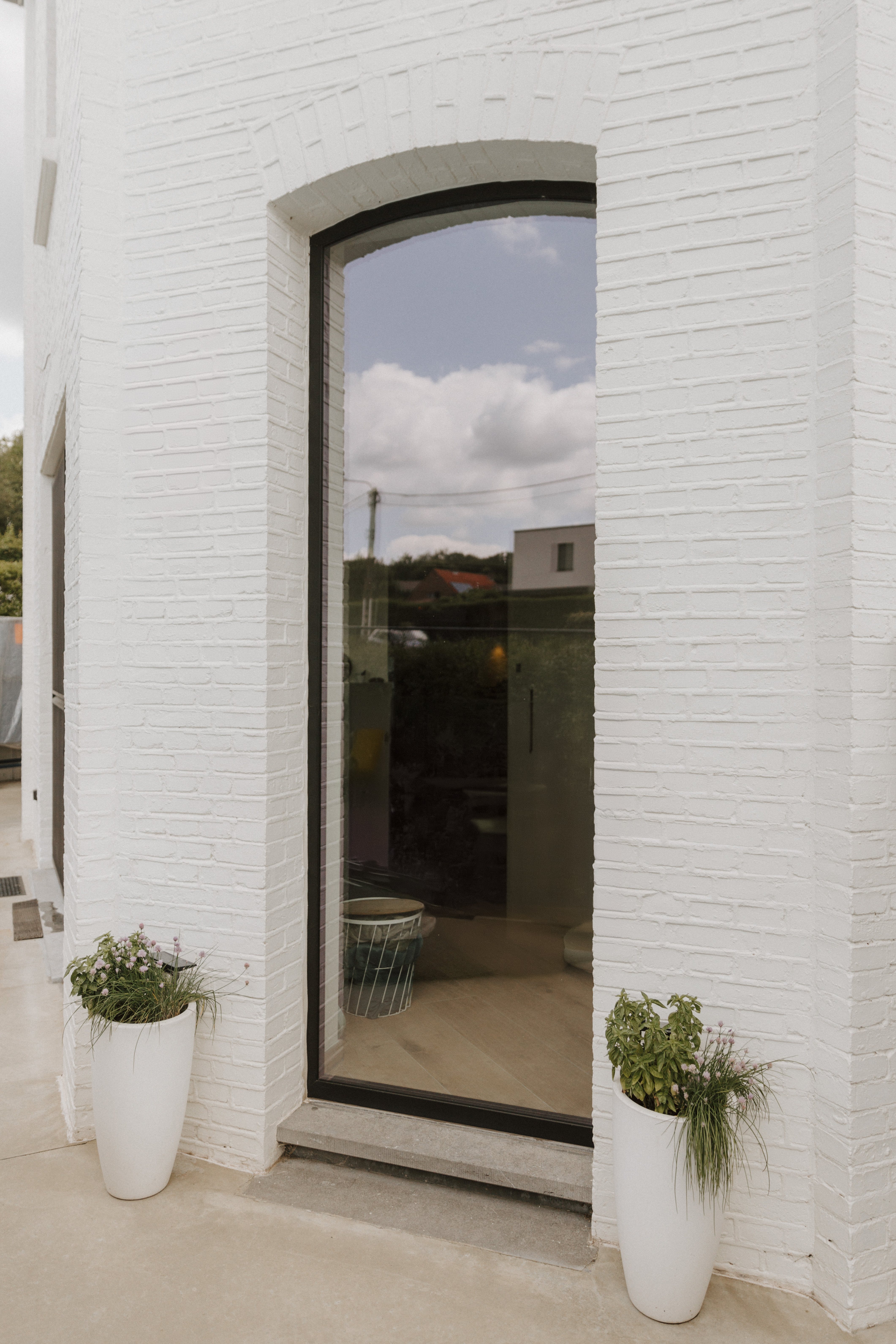 Hoog vast raam met diepzwart raamprofiel en afgeronde bovenzijde in een witte bakstenen gevel, met plantenpotten aan de voet.