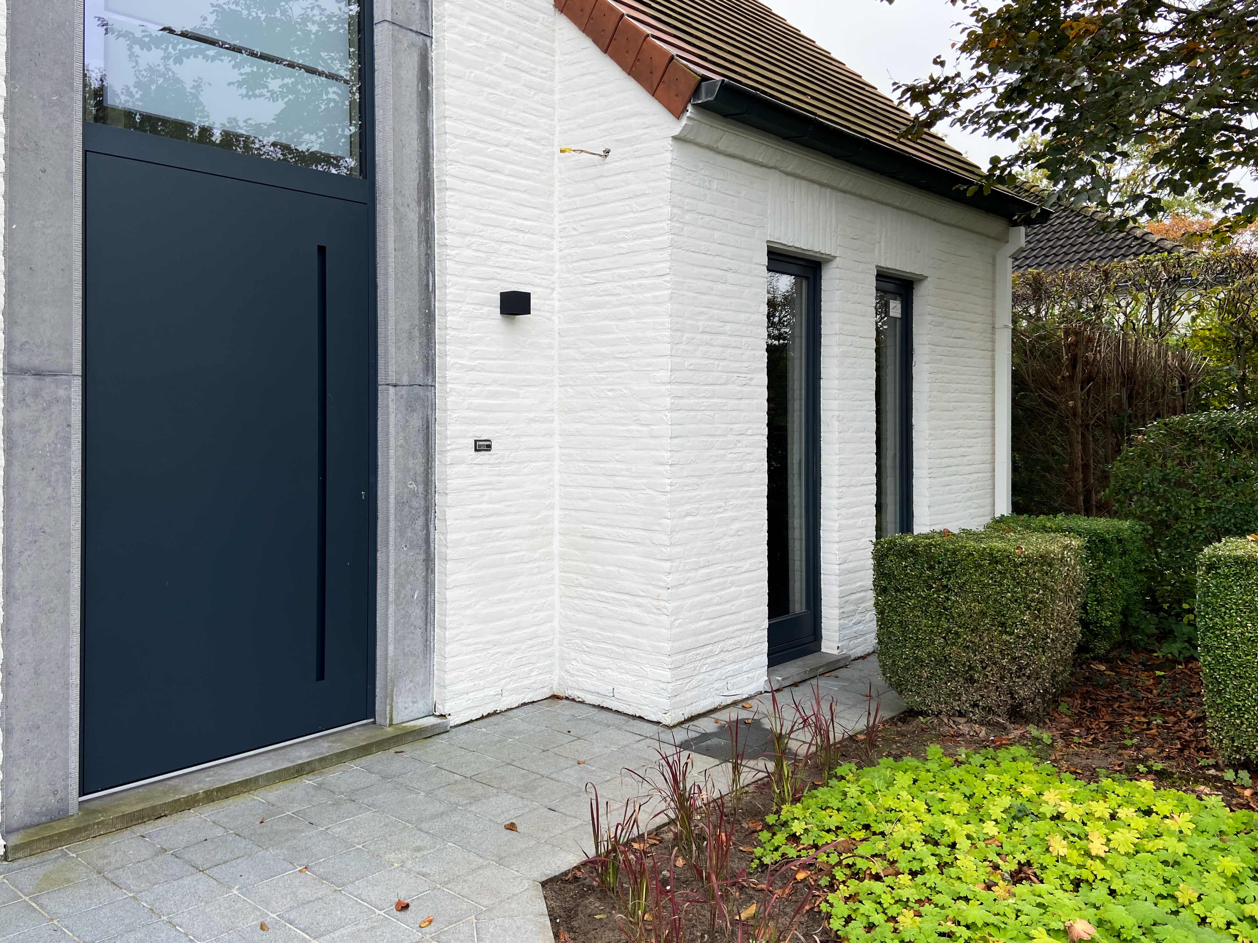Entrée d’une maison quatre façades avec façade en briques blanches, porte vitrée anthracite et deux fenêtres étroites, entourée de haies.