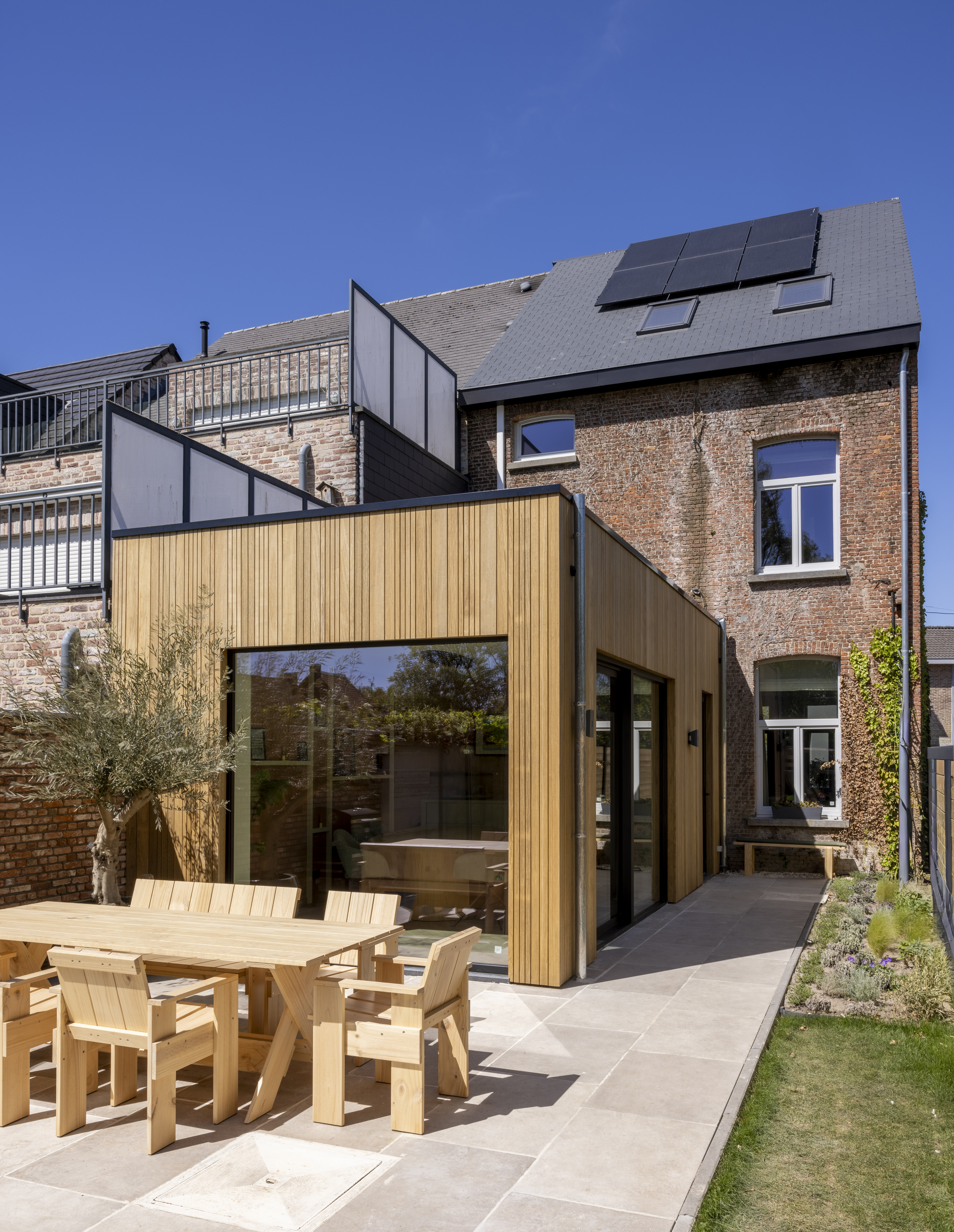 Moderne woninguitbreiding met een groot vast raam in signaalzwarte raamprofielen, naast een bakstenen gevel en een terras met houten tuinmeubilair.