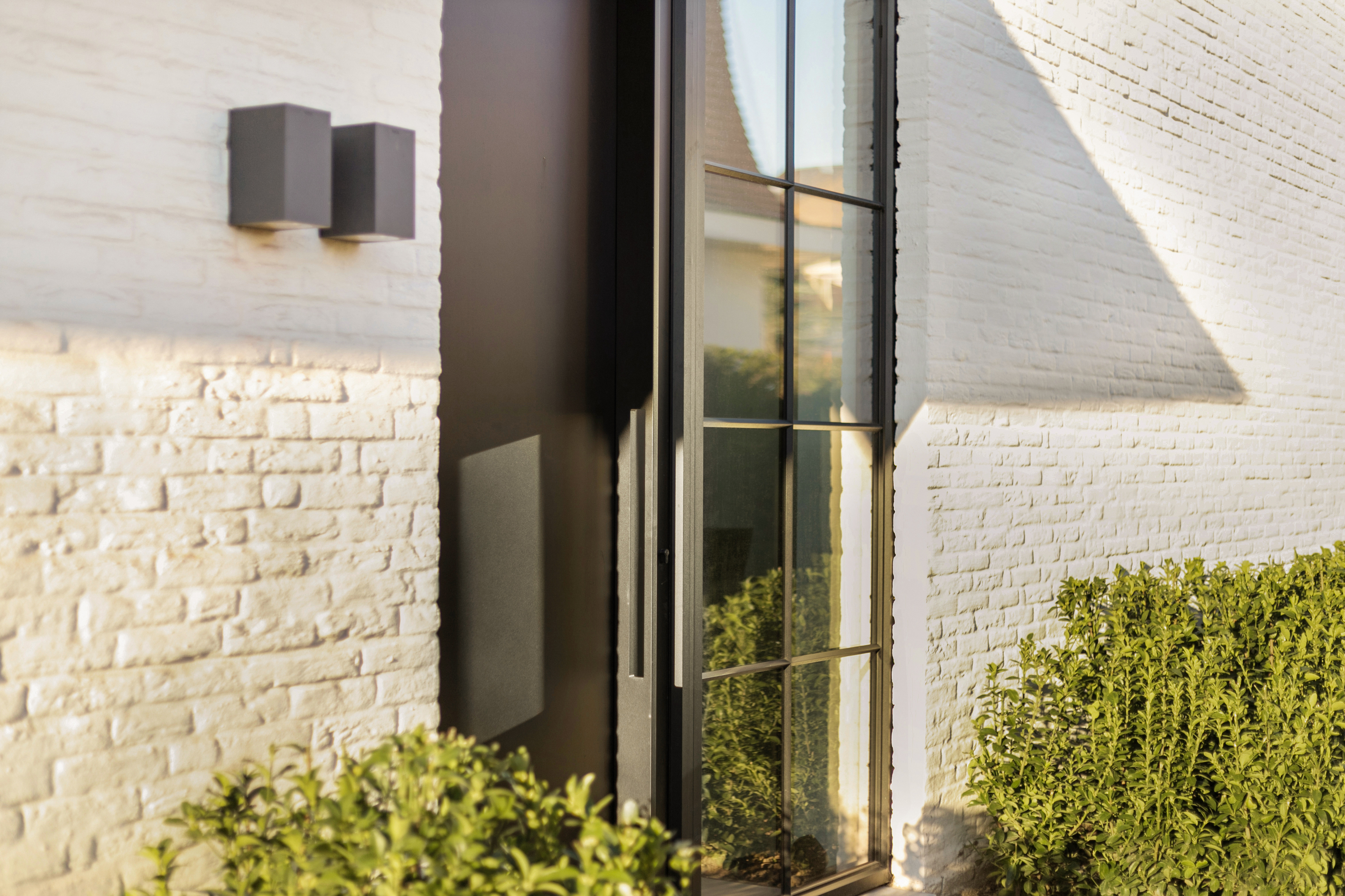 Hoog vast raam in Steellook-stijl met zwarte aluminiumprofielen in een witte bakstenen gevel, naast een inkom en omringd door groene struiken.