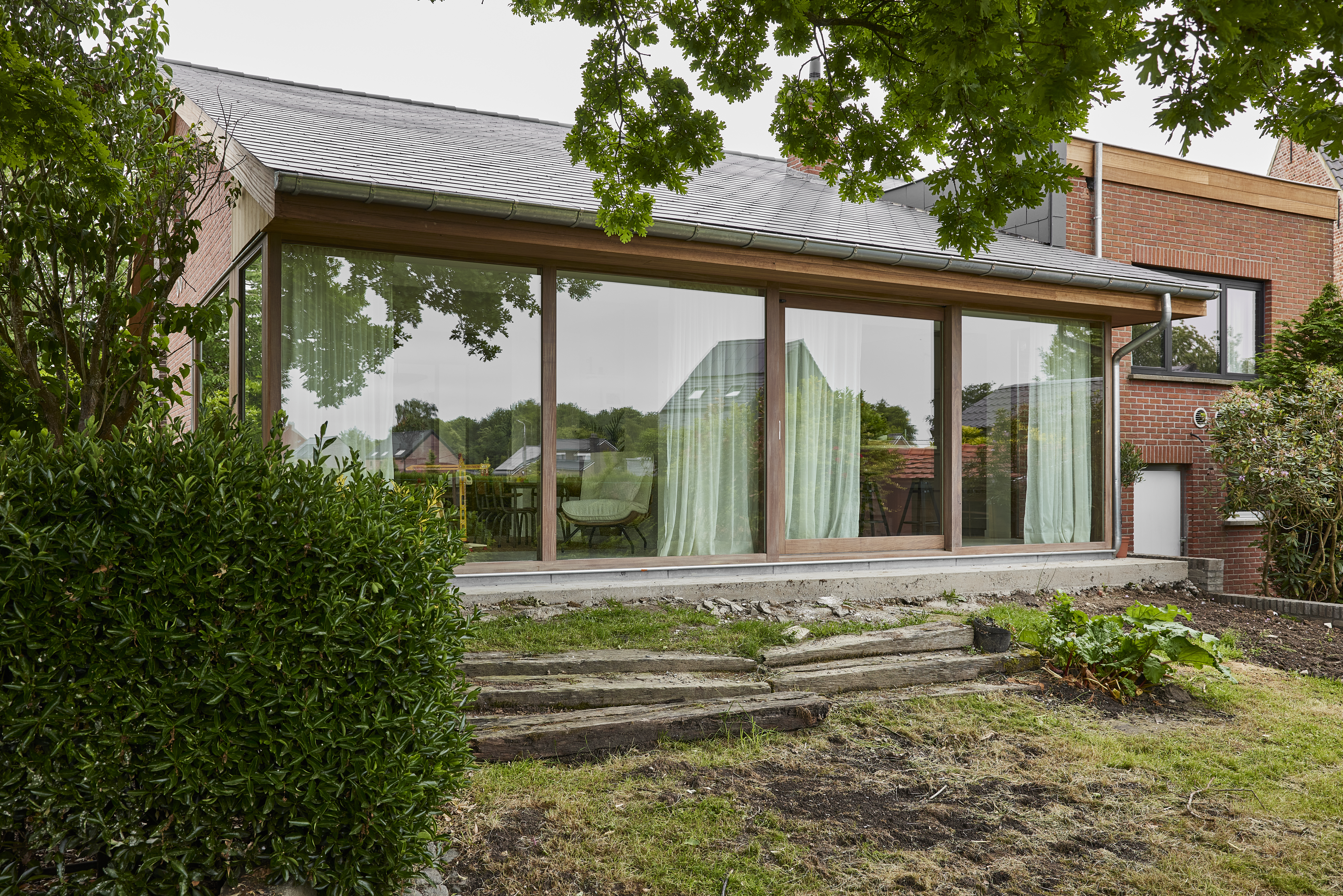 Glazen tuingevel met raamprofielen in Afromosia-hout met Rubio Natural-afwerking, aansluitend op een bakstenen woning en een aangelegde tuin.