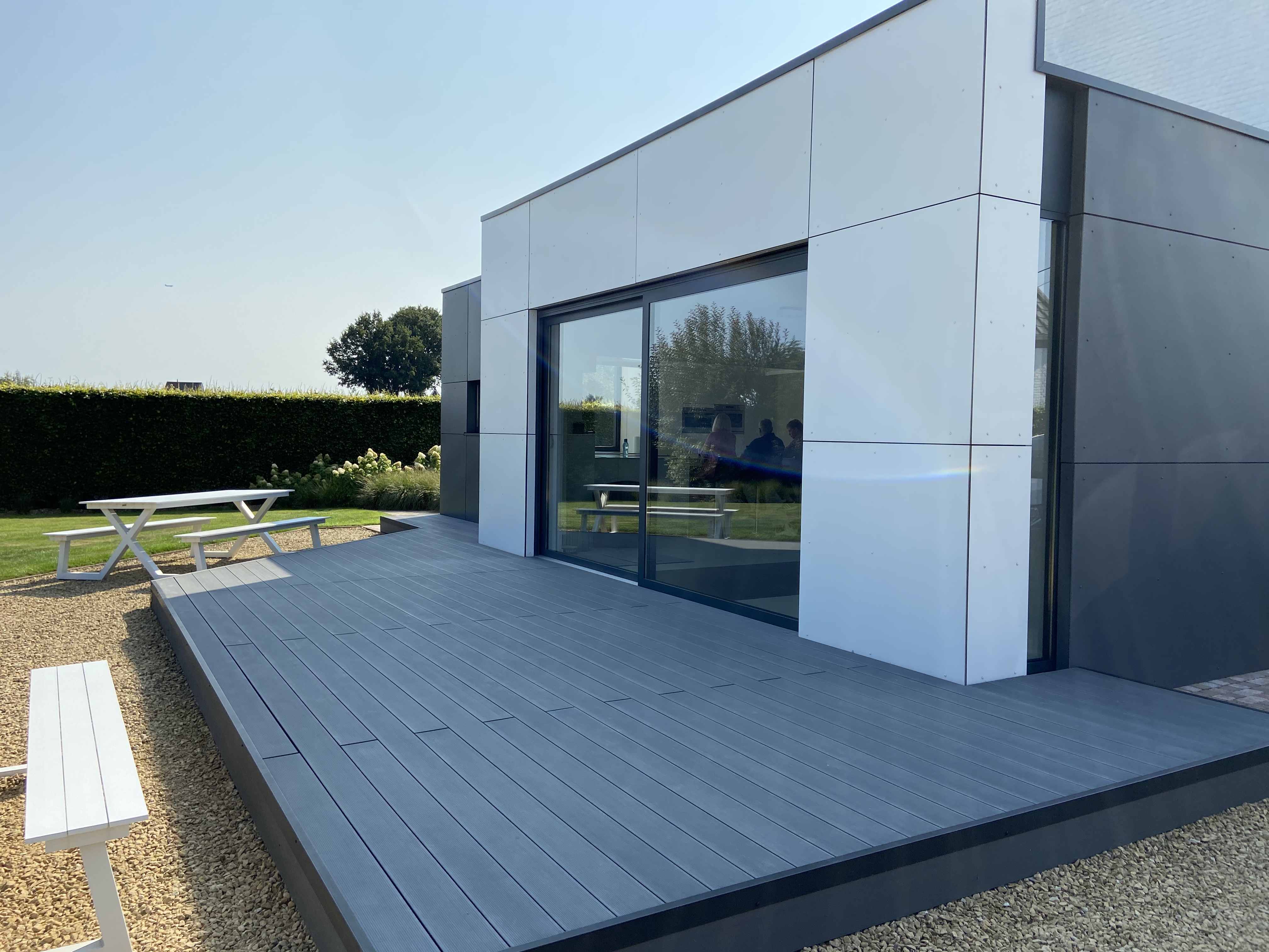 Moderne vrijstaande woning met grote donkere glaspartij aan een ruim grijs terras, naast een grasveld met houten picknicktafel.