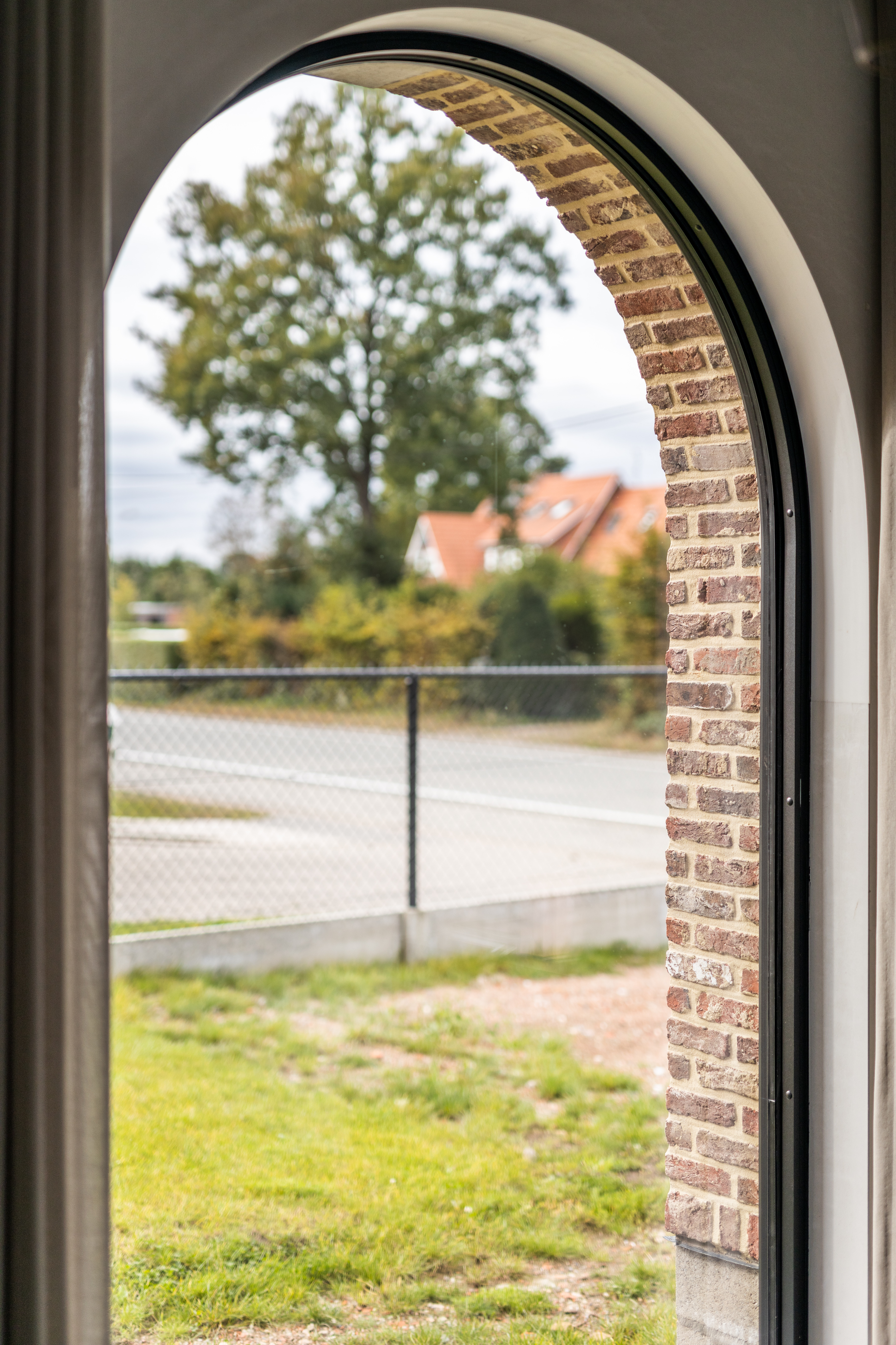 Zwart vast raam met ronde boog in een bakstenen muur, met zicht op de tuin, omheining, straat en landelijke woningen.
