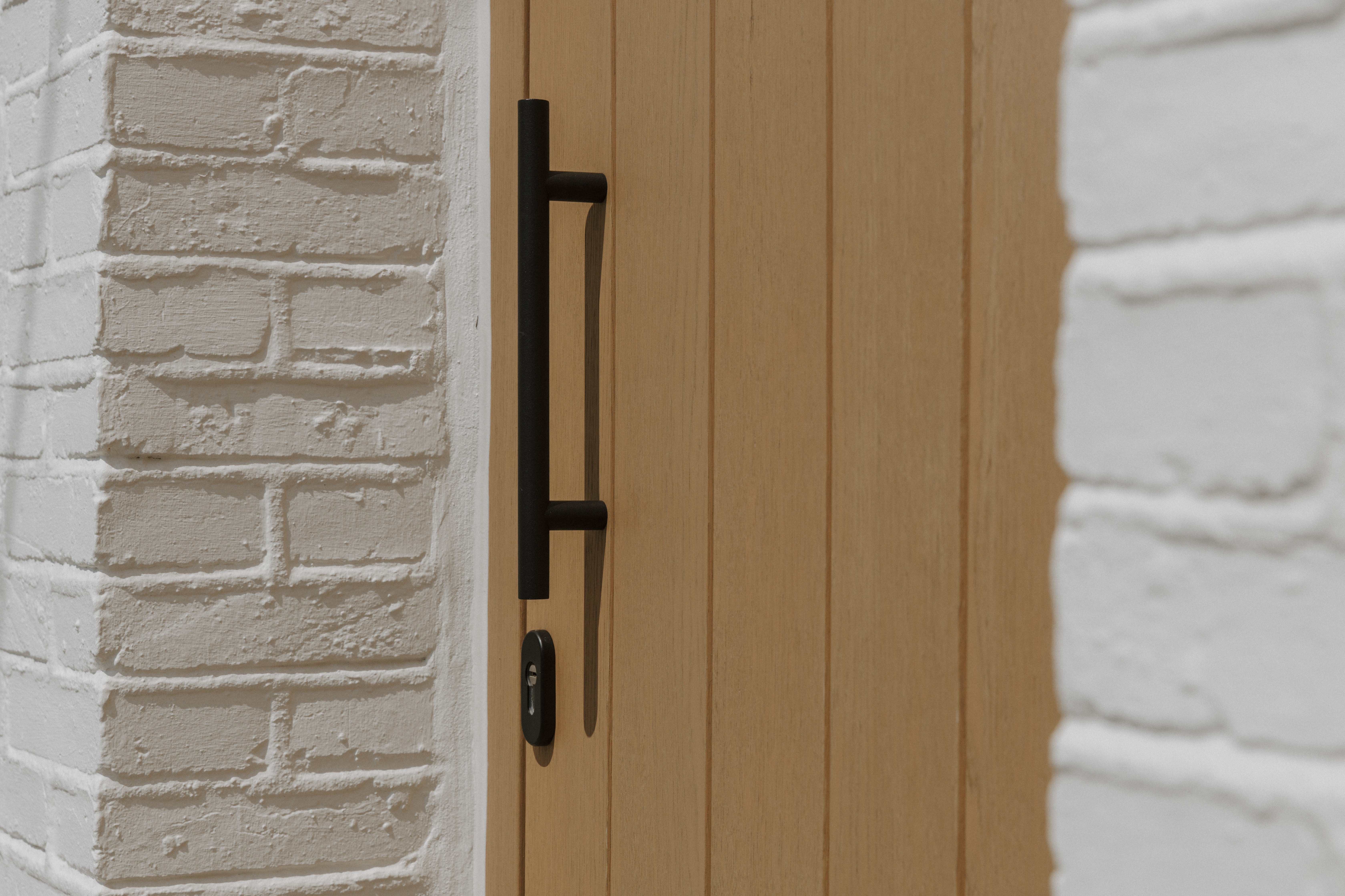 Close-up van een eikenhouten deur met Rubio Natural-afwerking, een zwarte verticale deurklink en slotcilinder tussen witte bakstenen muren.