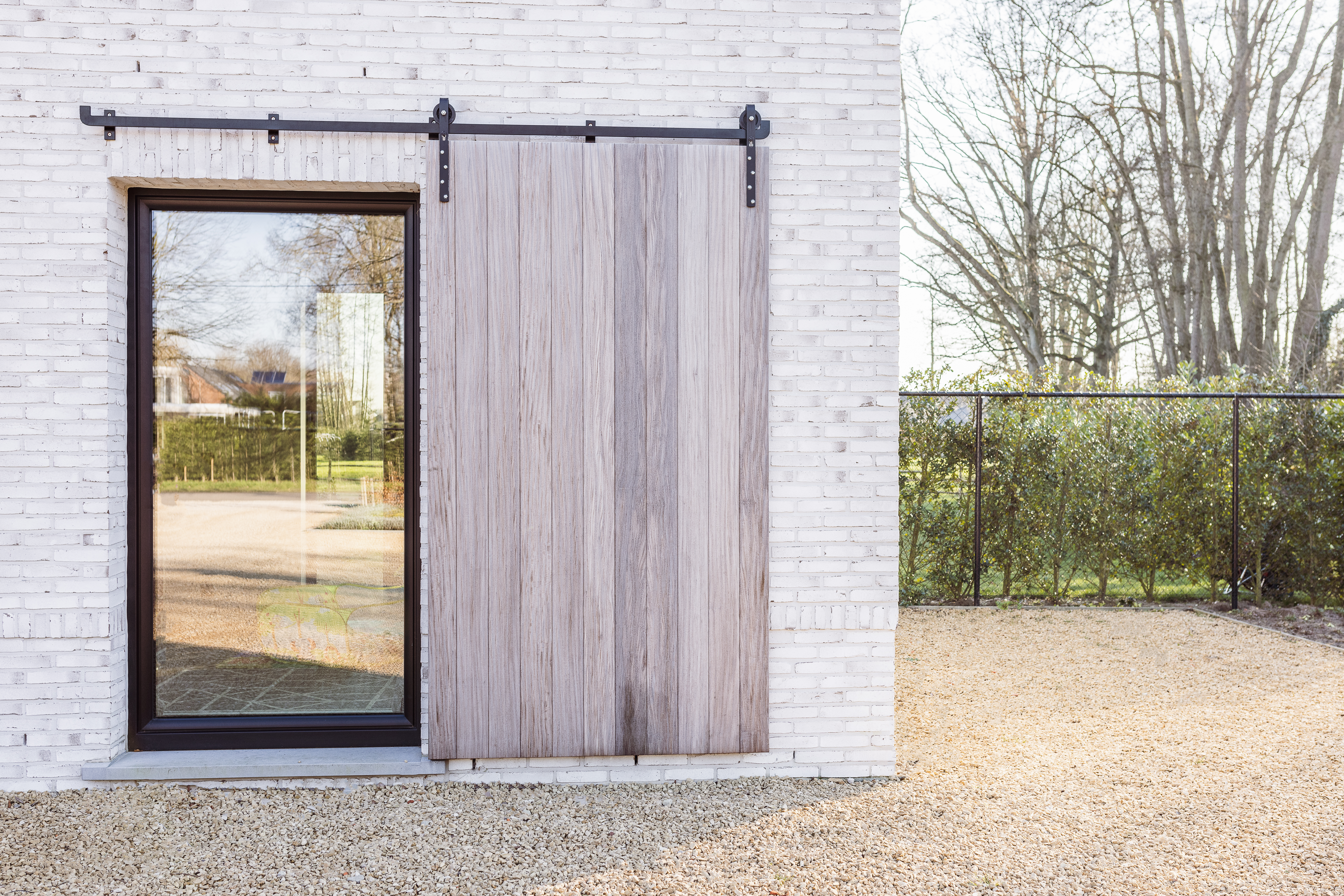 Mat zwart draai-kipraam met ronde Impakt PLUS-beslagonderdelen naast een houten schuifluik aan een witte bakstenen gevel.