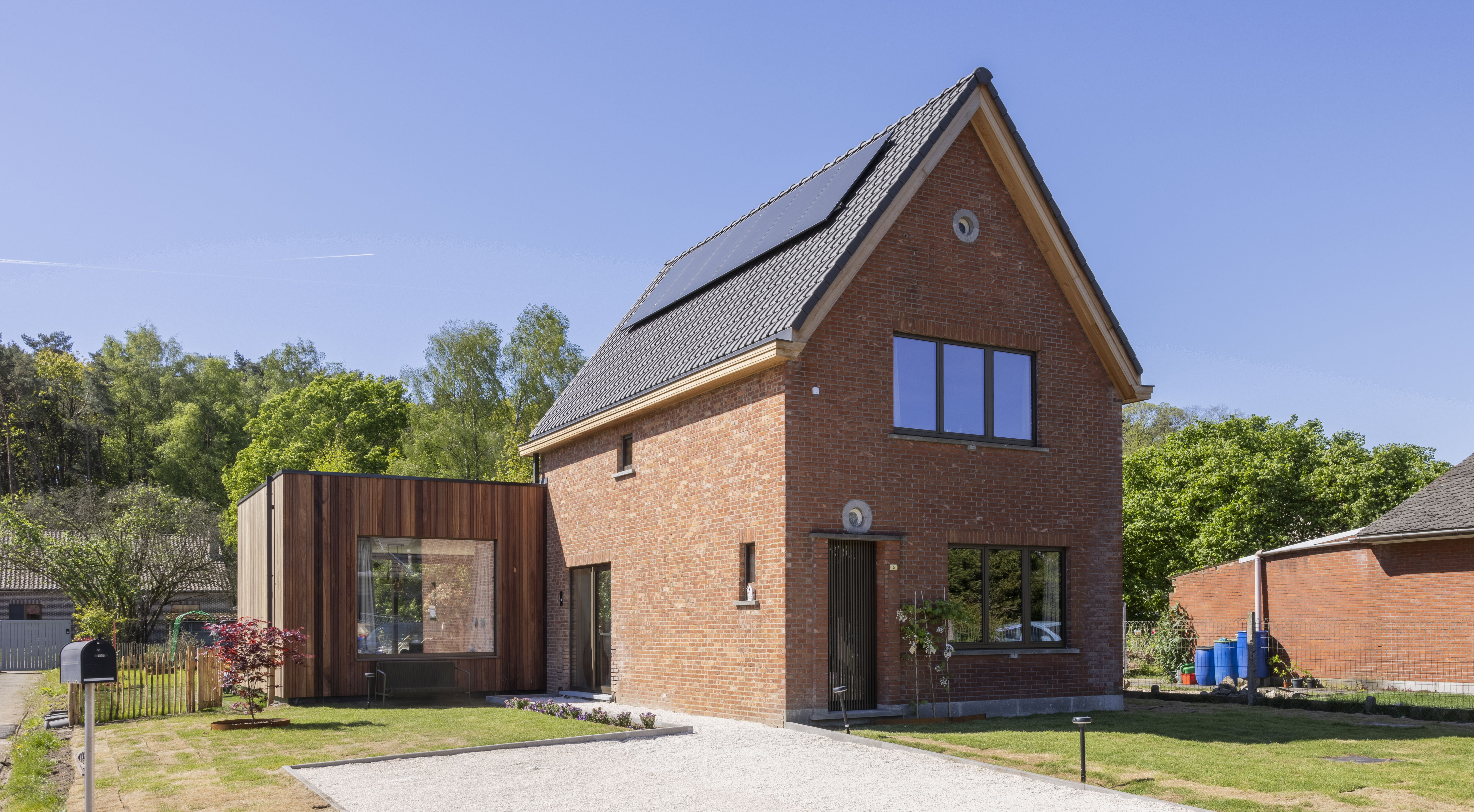 Moderne bakstenen woning met een driedubbel raam en vast raam in donkere raamprofielen, naast een houten aanbouw en verzorgde tuin.