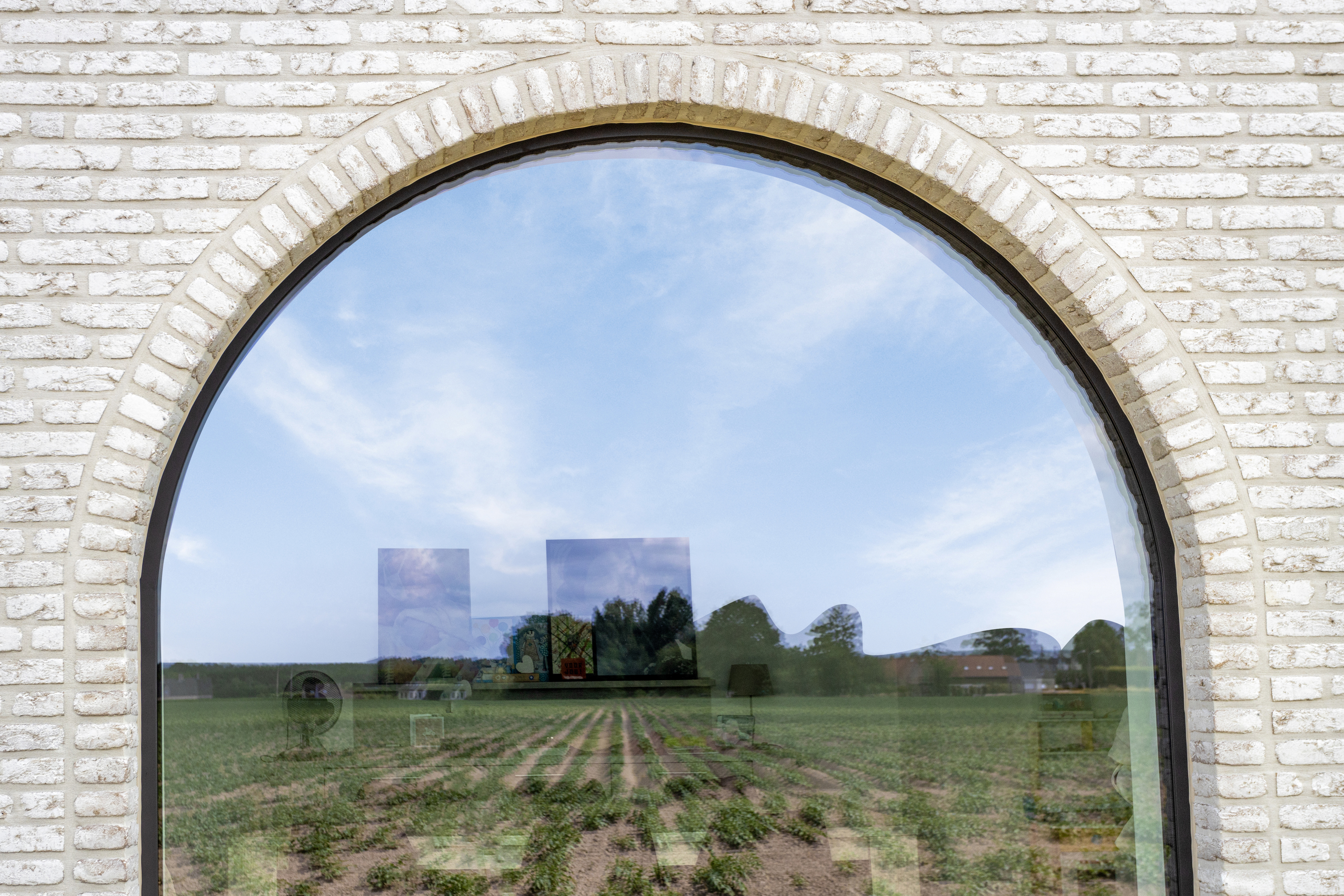 Vast raam in signaalzwart met boogvormige bovenkant, ingebouwd in een lichte bakstenen gevel van een landelijke nieuwbouwwoning met zicht op velden.
