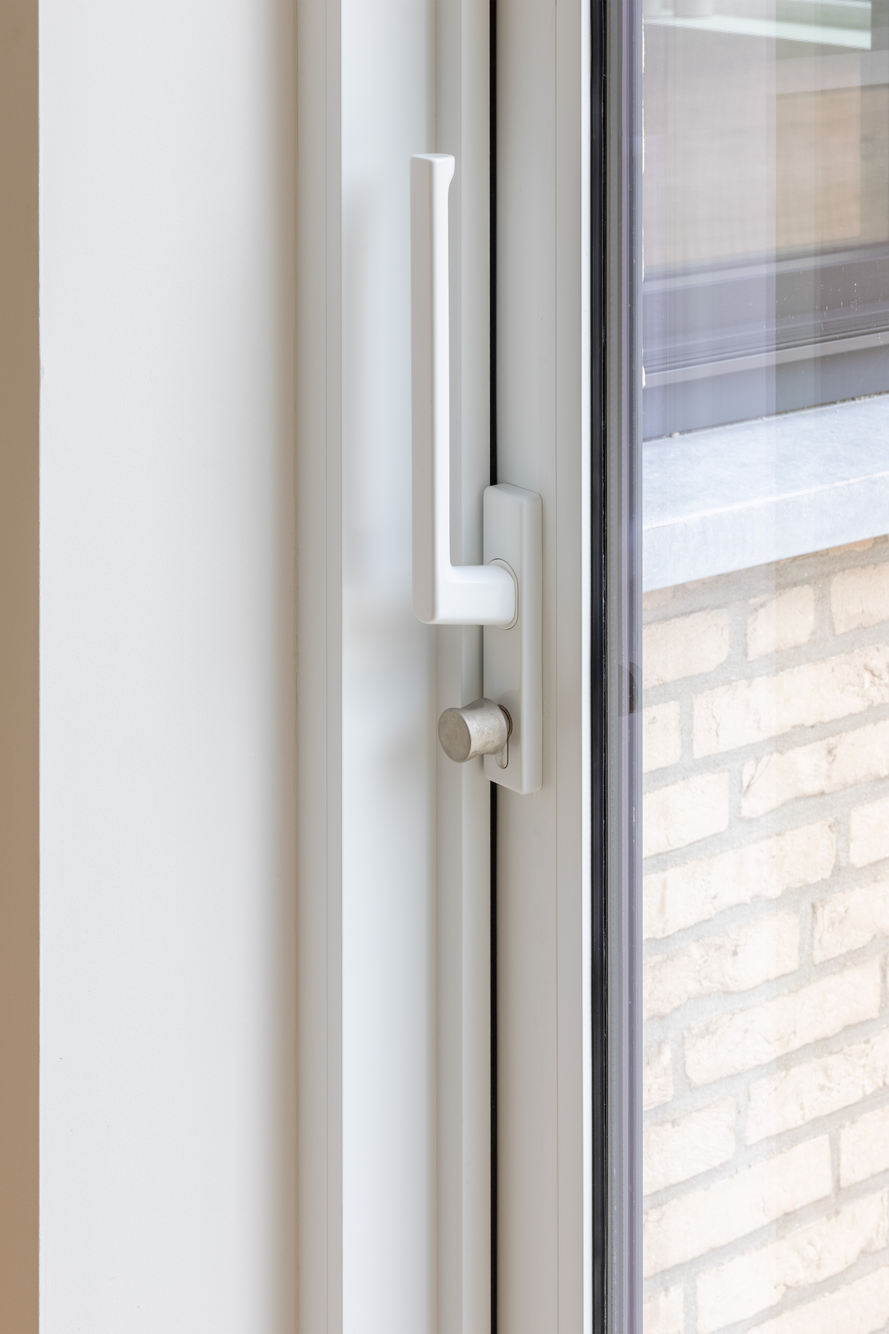 Close-up van een matwit aluminium schuifraam met verticale handgreep en sluiting, naast een lichte bakstenen gevel in een gesloten bebouwing.