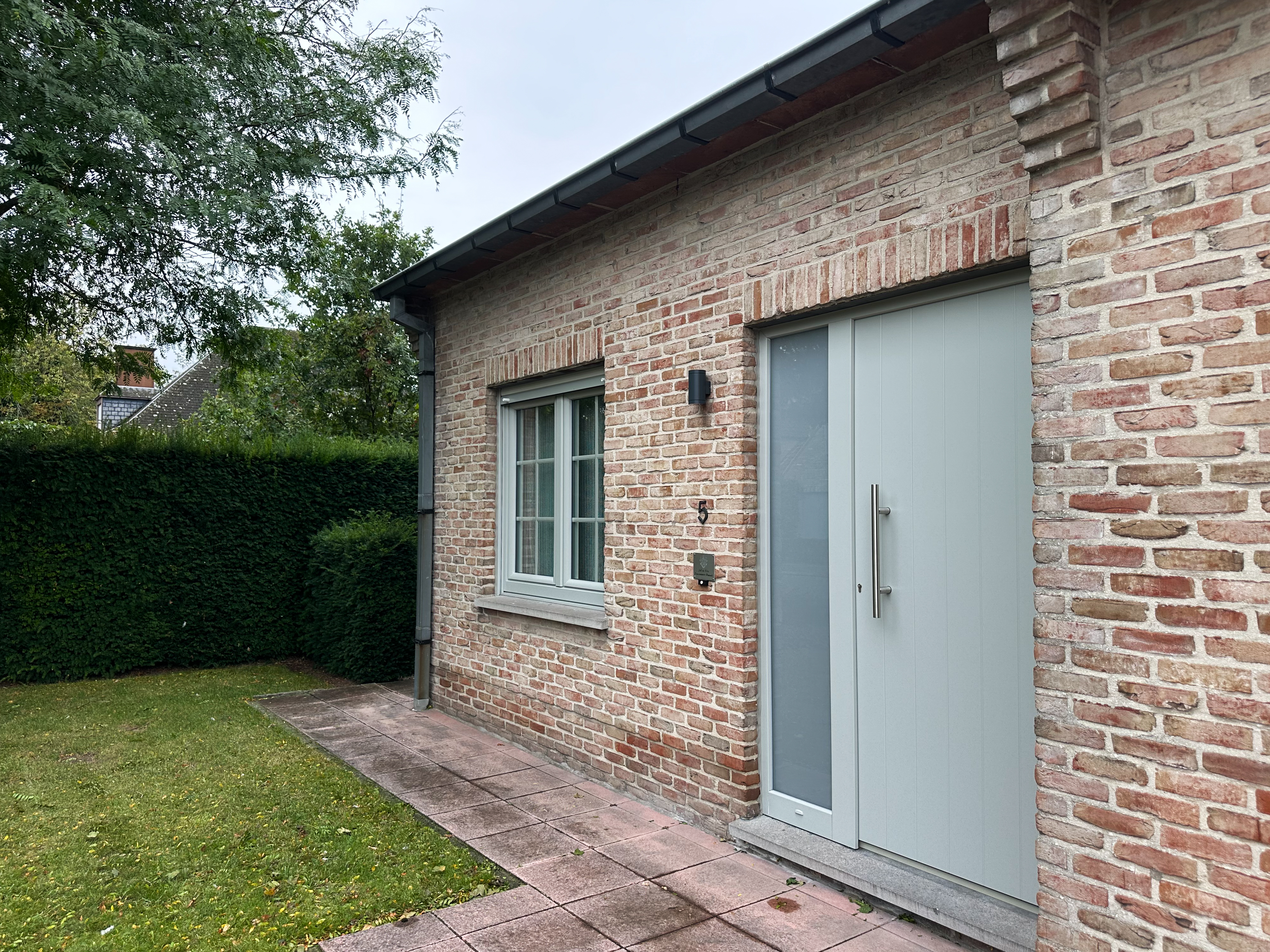 Agaatgrijs PVC-draai-kipraam en deur met zijlicht en paneel in een gemetselde gevel, naast een verzorgde tuin.