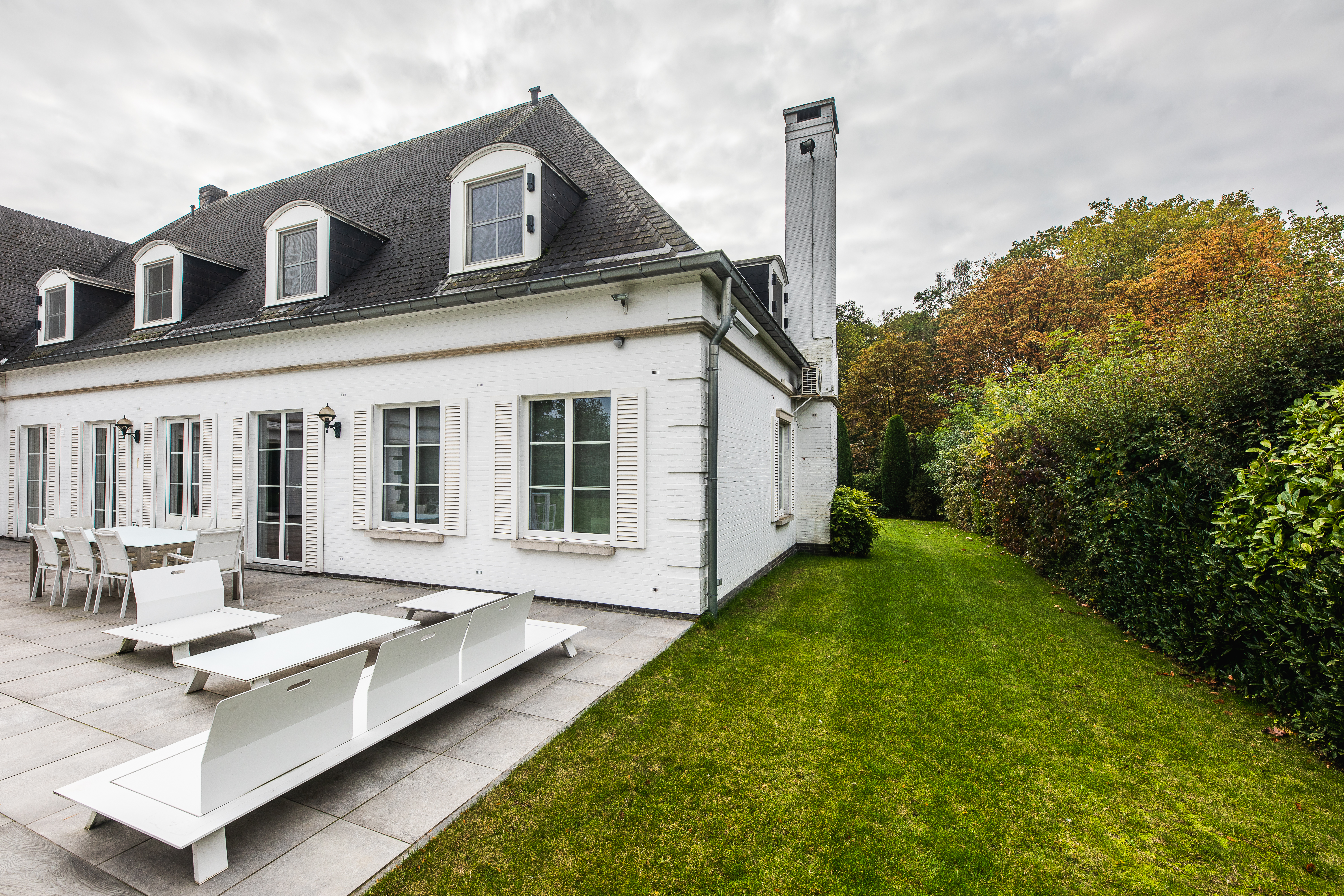 Witte vrijstaande woning met verfijnde klassieke gevel, dakramen en grote ramen, met een ruim terras en een verzorgde tuin.