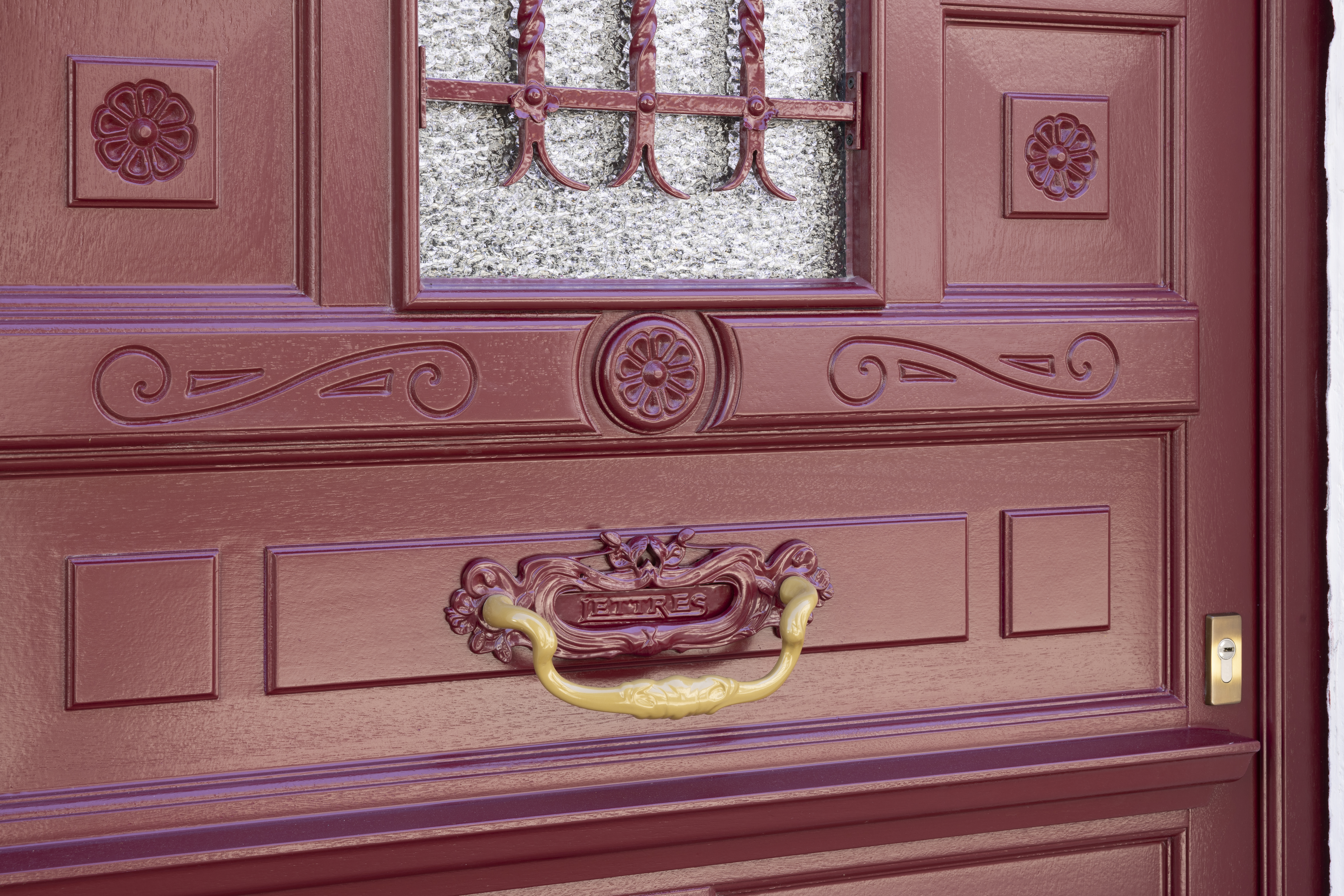 Close-up van een wijnrode Meranti-houten deur met decoratieve panelen, sierdetails, een kunstige deurklink en een glazen bovenlicht.