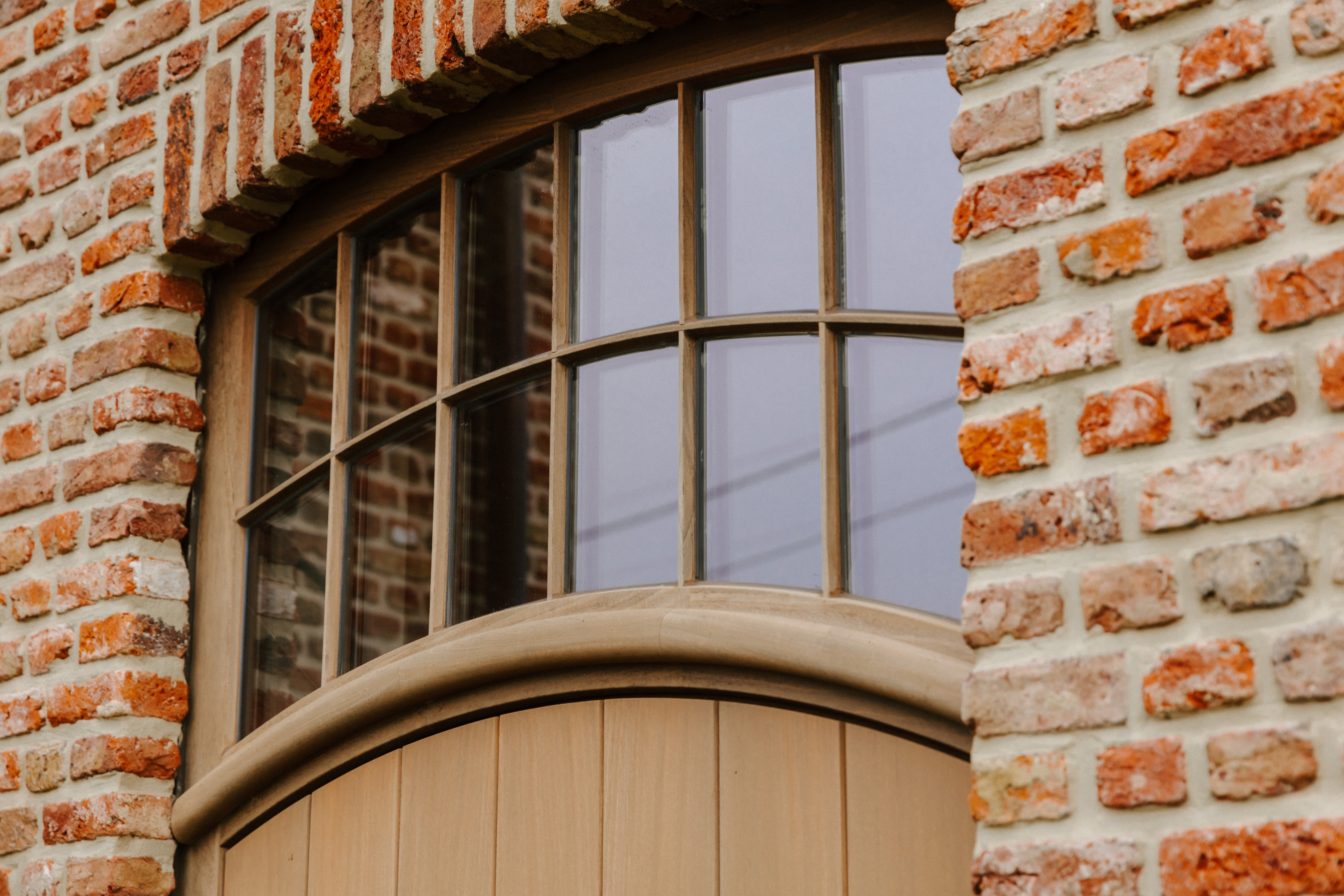 Detail van een warme, landelijke bakstenen gevel met een Afrormosia-houten deur in Rubio Natural en een gebogen bovenlicht bij een nieuwbouw.