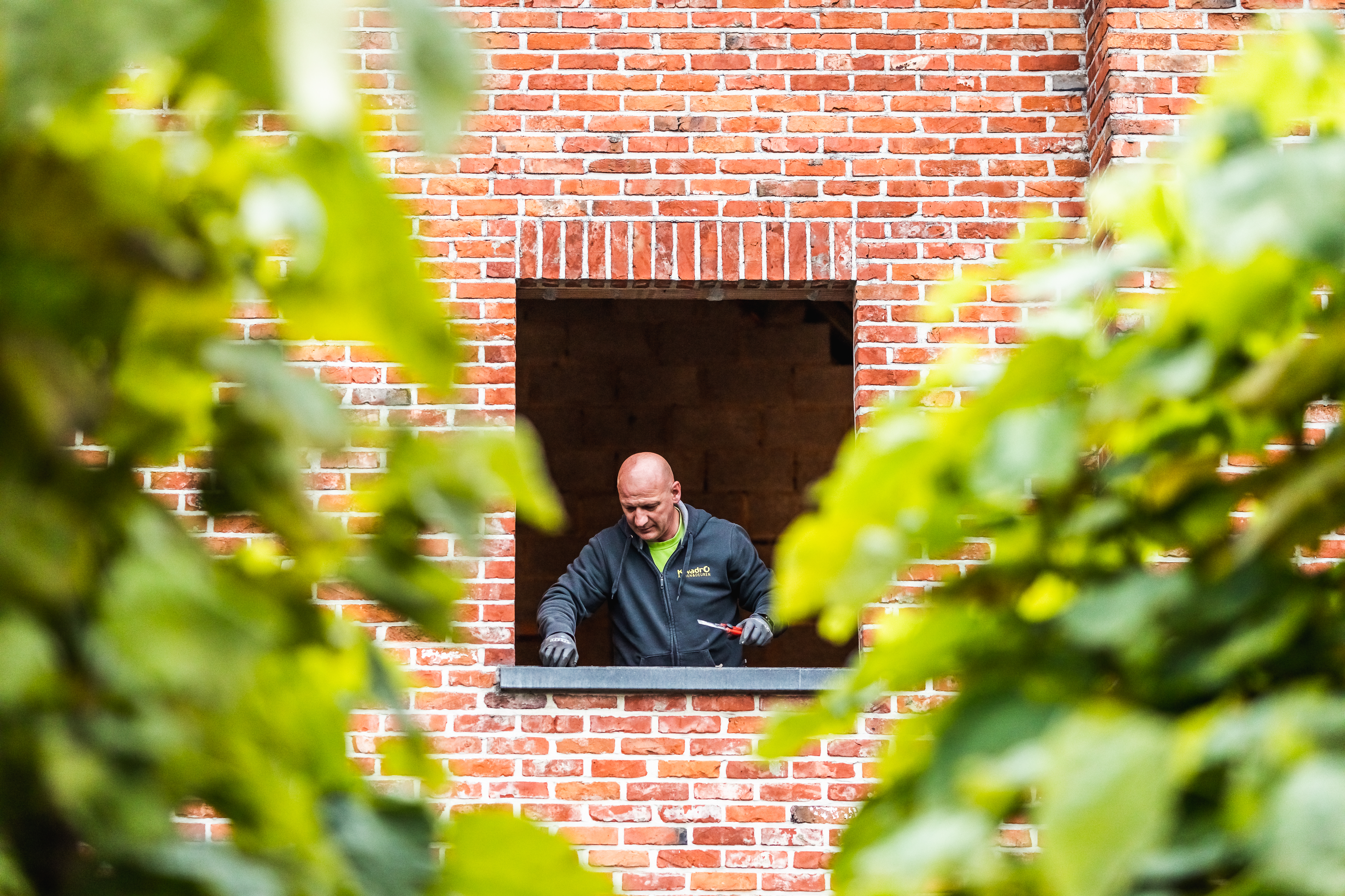 Een medewerker kijkt door een uitsparing in een gevel van rode baksteen, omringd door groene bladeren op de voorgrond.