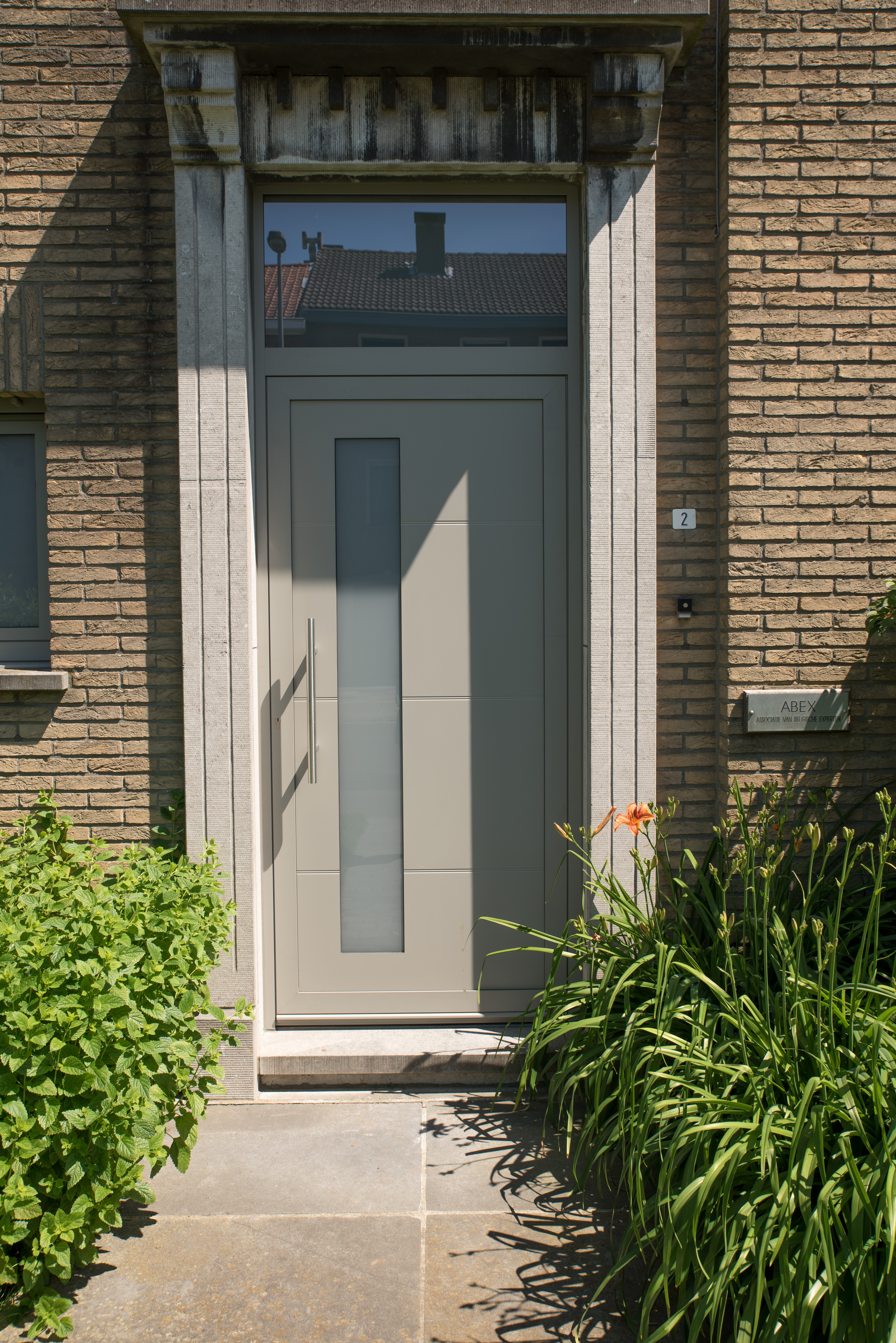 Lichtgrijze semi-beglaasde voordeur in de bakstenen gevel van een gerenoveerde driegevelwoning, met een groen aangelegde toegang.