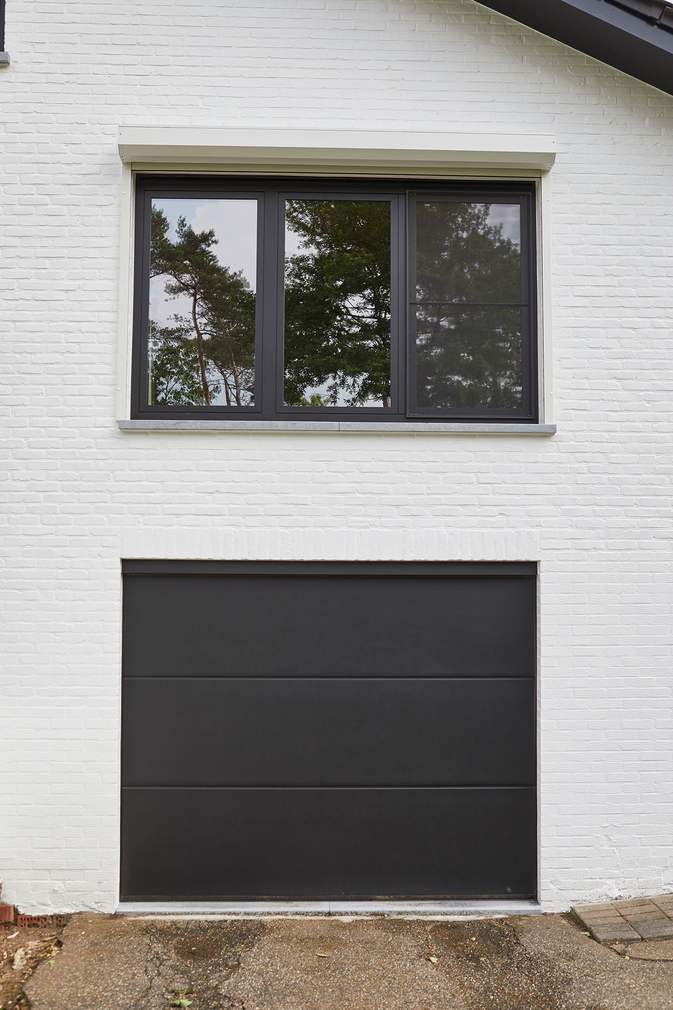 Zwart steellook raam met driedubbele beglazing boven een moderne zwarte garagepoort in de witte gevel van een gerenoveerde open bebouwing.