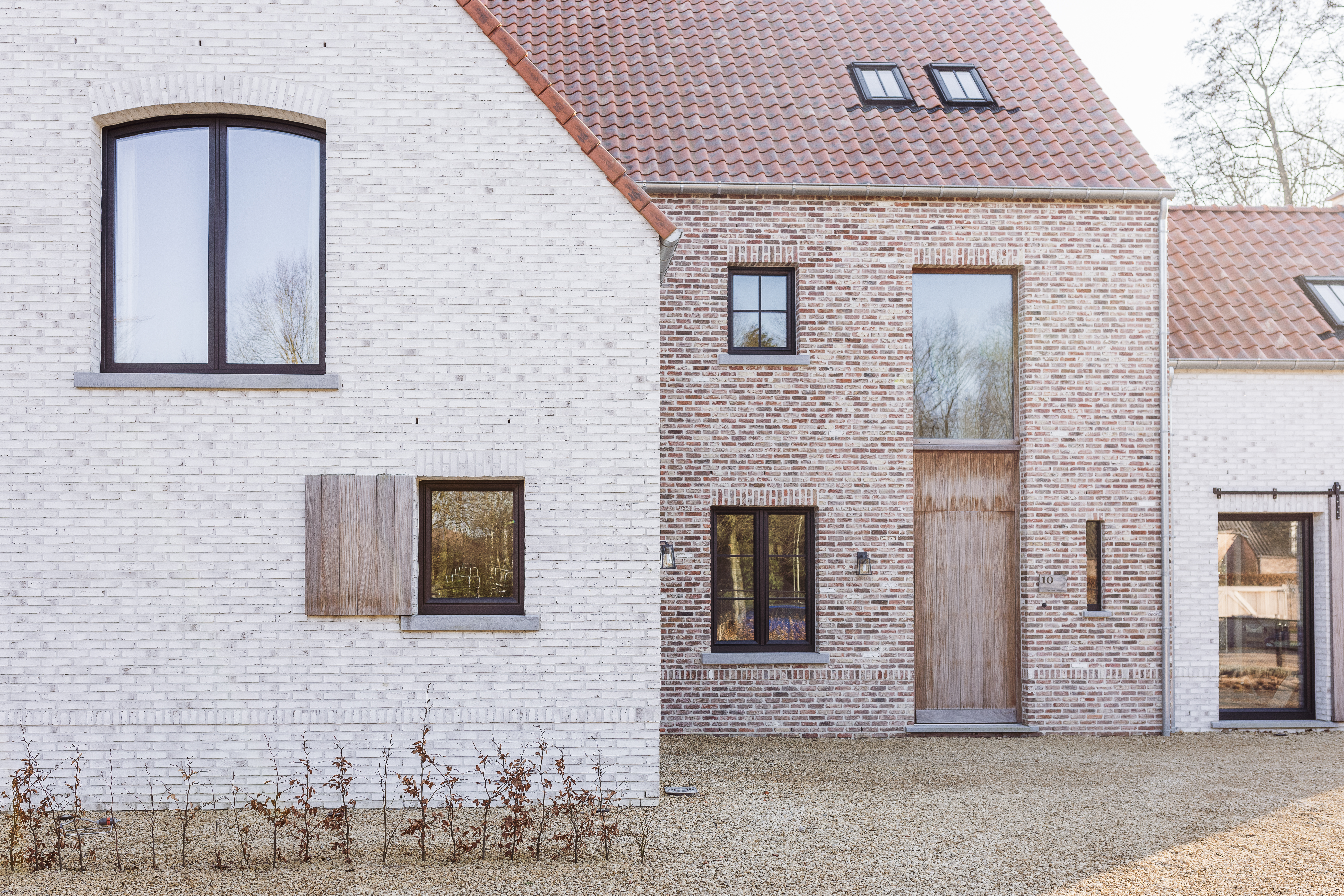 Gevel van een woning in lichte en rode baksteen, met matzwarte ramen, waaronder een dubbel draai-kipraam, en een hoge houten deur.