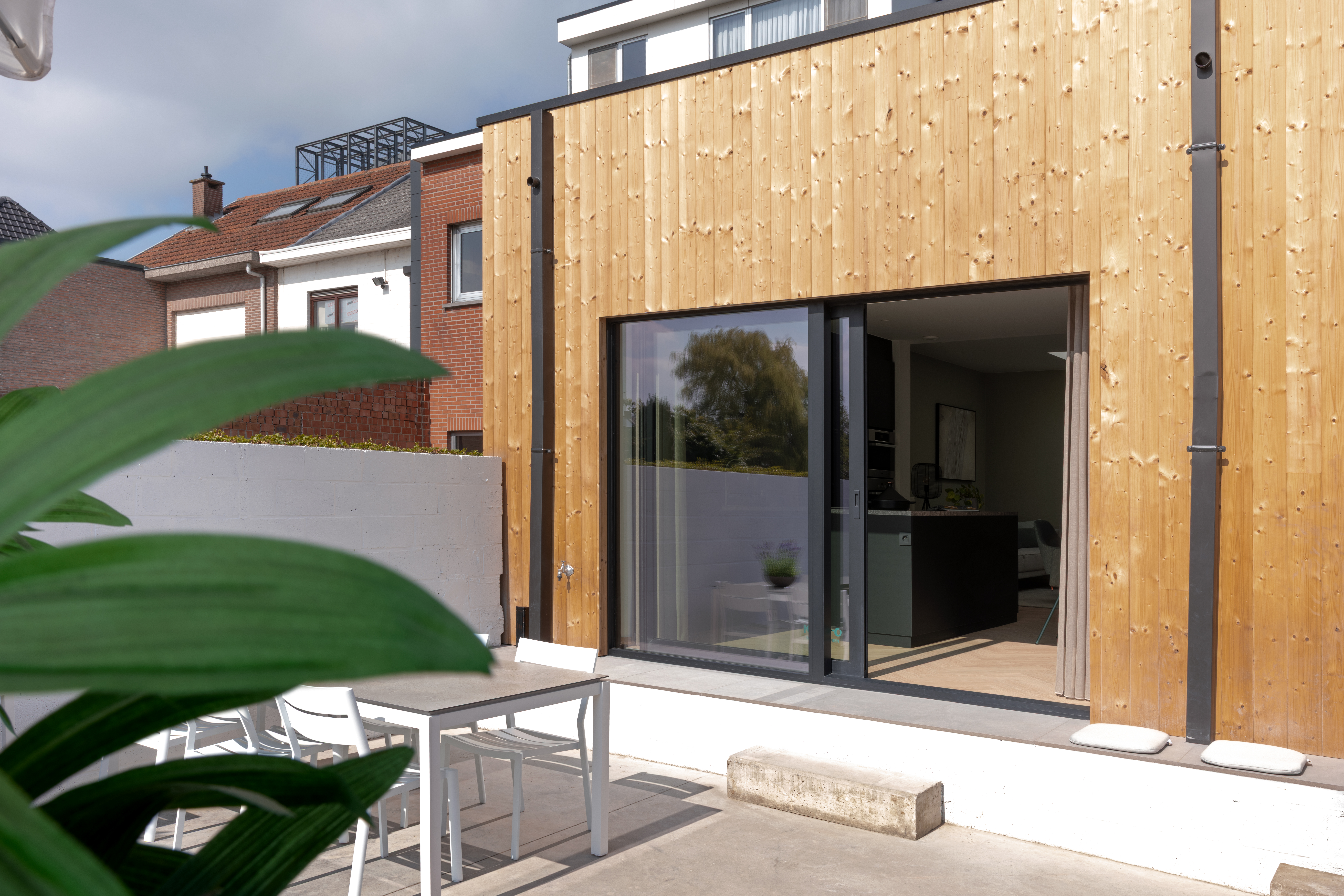Moderne aanbouw met verticale houten gevelbekleding en een groot zwart schuifraam naar een terras met tafel en stoelen.