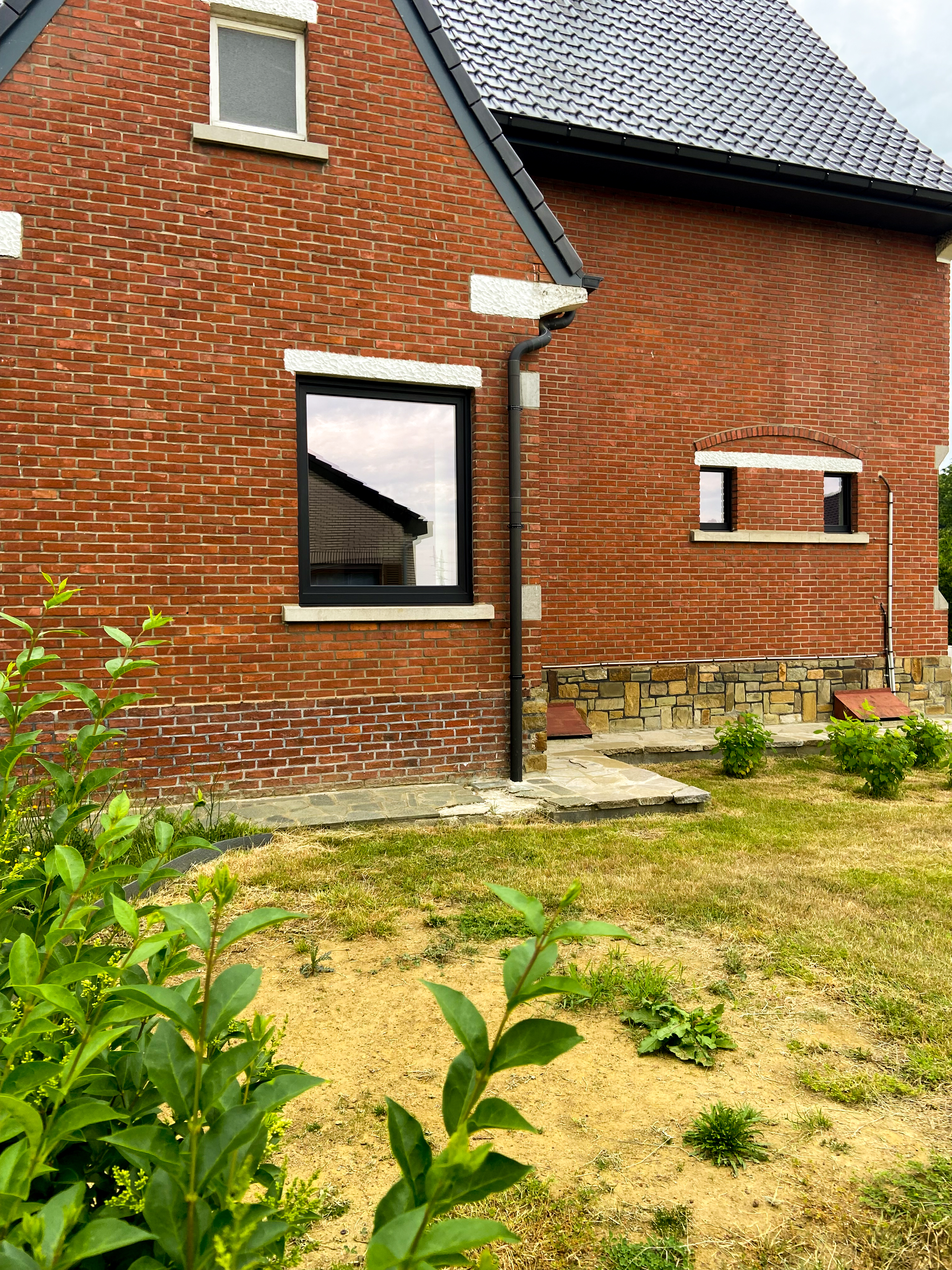 Gerenoveerde vrijstaande woning met een rood bakstenen gevel, matzwarte raamprofielen en een klein raam in de zijgevel, omgeven door een tuin.
