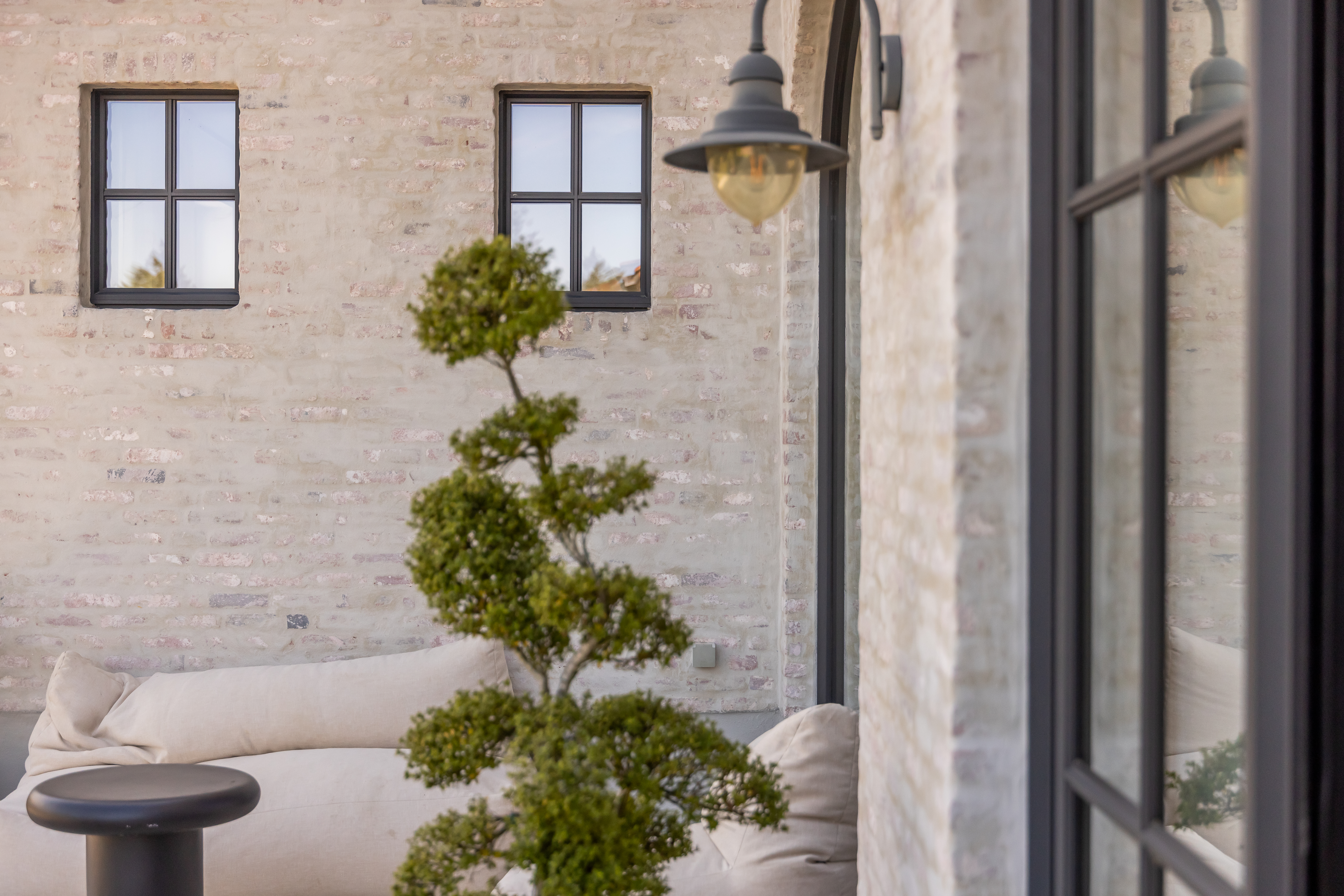 Zwarte raamprofielen in steellook met een vast raam en draai-kipraam in een warm, landelijk interieur met bakstenen muur, sofa en vormboom.