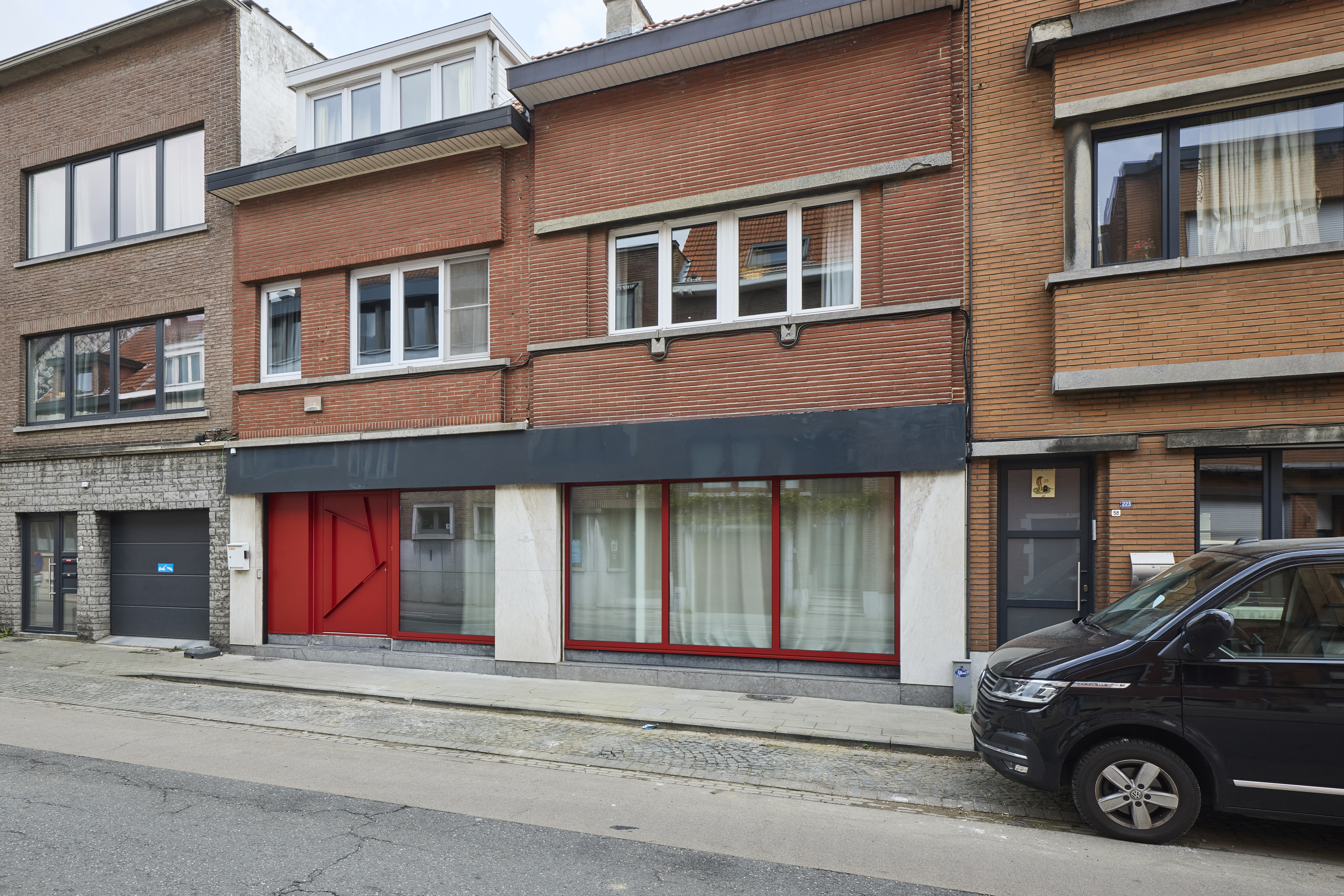 Straatgevel met een groot vast ALU-Komfort EVO-raam met driedubbele beglazing en rode raamprofielen, geïntegreerd in de benedenverdieping van een bakstenen gebouw.