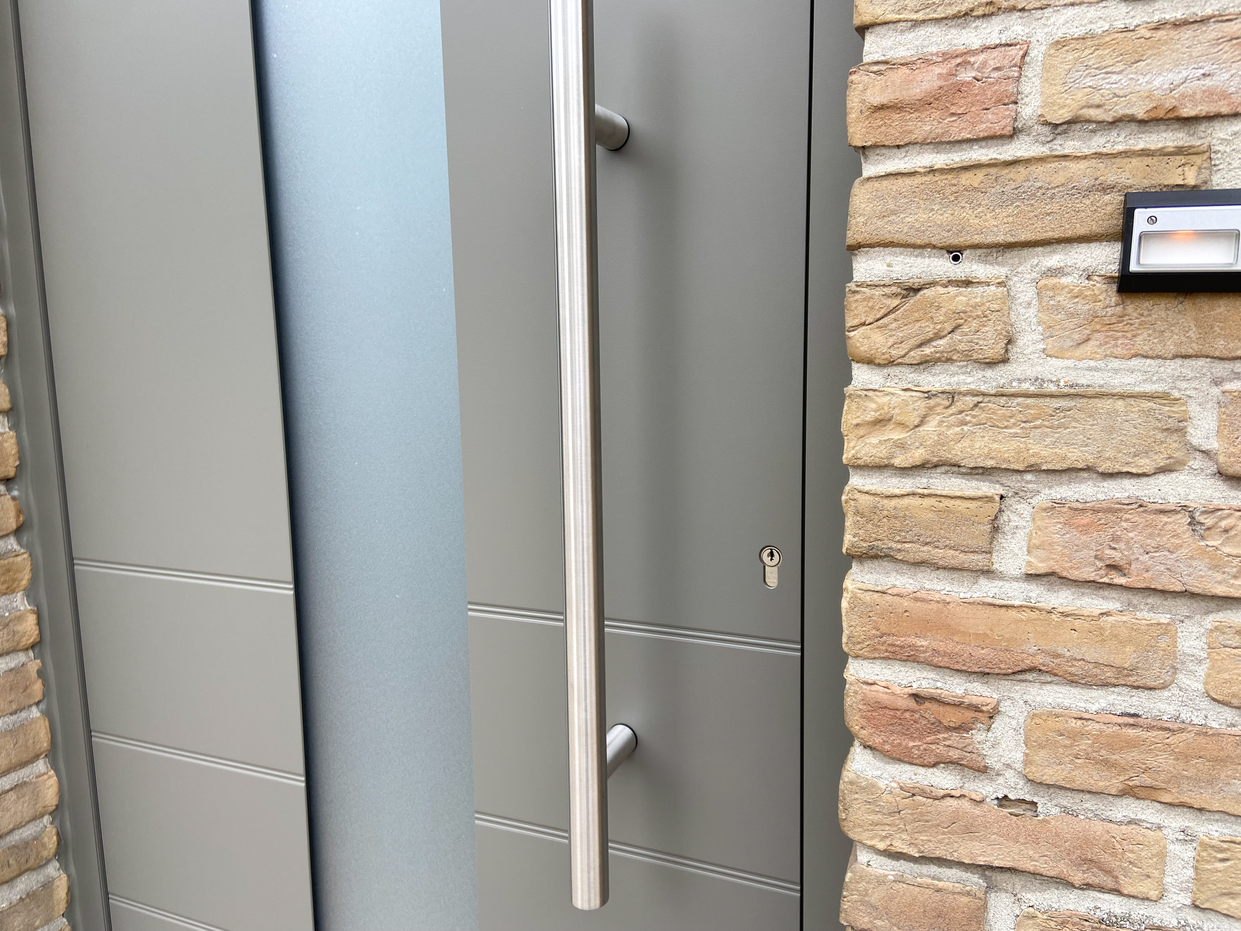 Grijze buitendeur met lange verticale handgreep en een zijpaneel in mat glas, verwerkt in een warme bakstenen gevel van een tijdloos gerenoveerde open bebouwing.