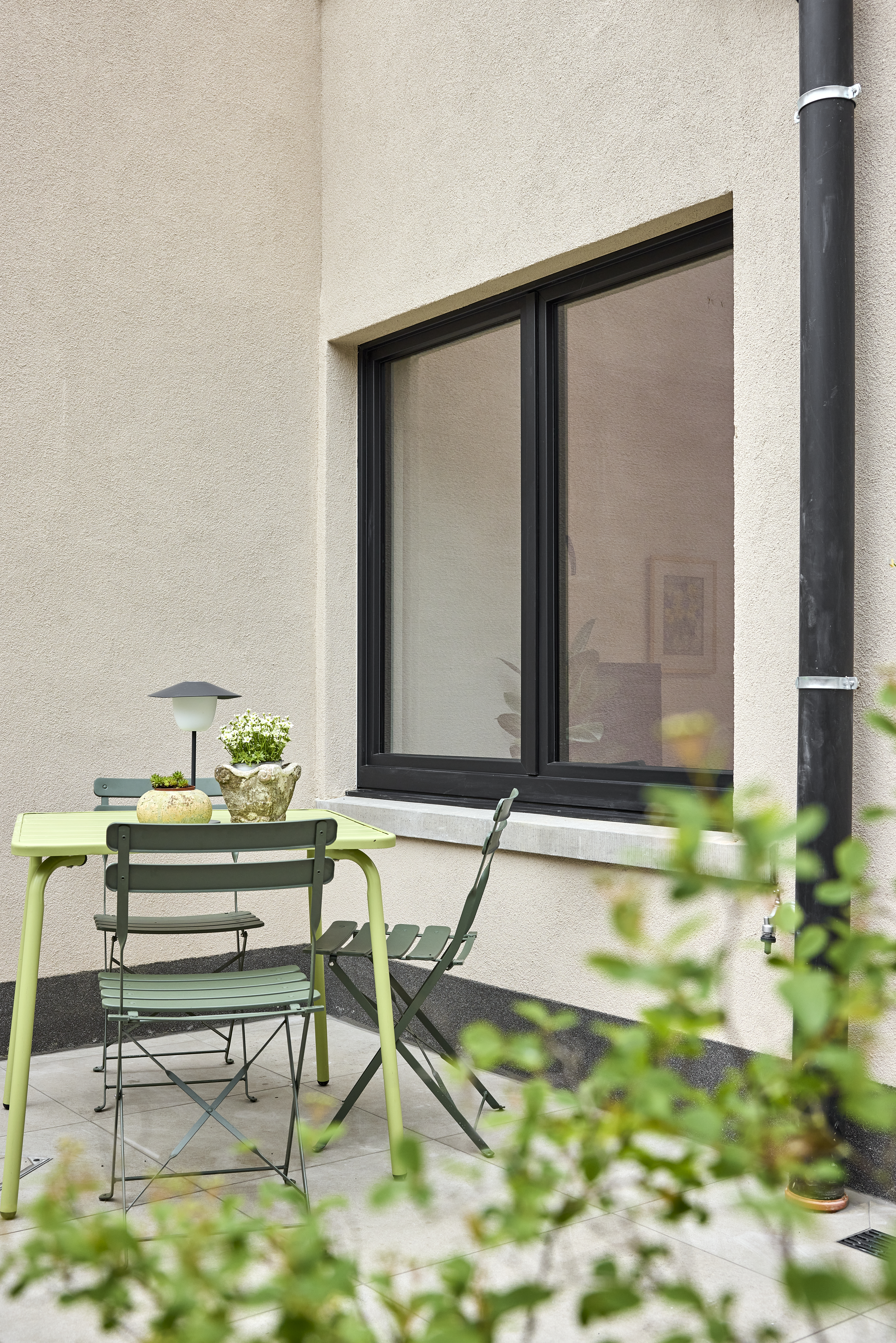Fenêtre double oscillo-battante noire mate dans une façade claire, à côté d’une petite terrasse avec une table et des chaises.
