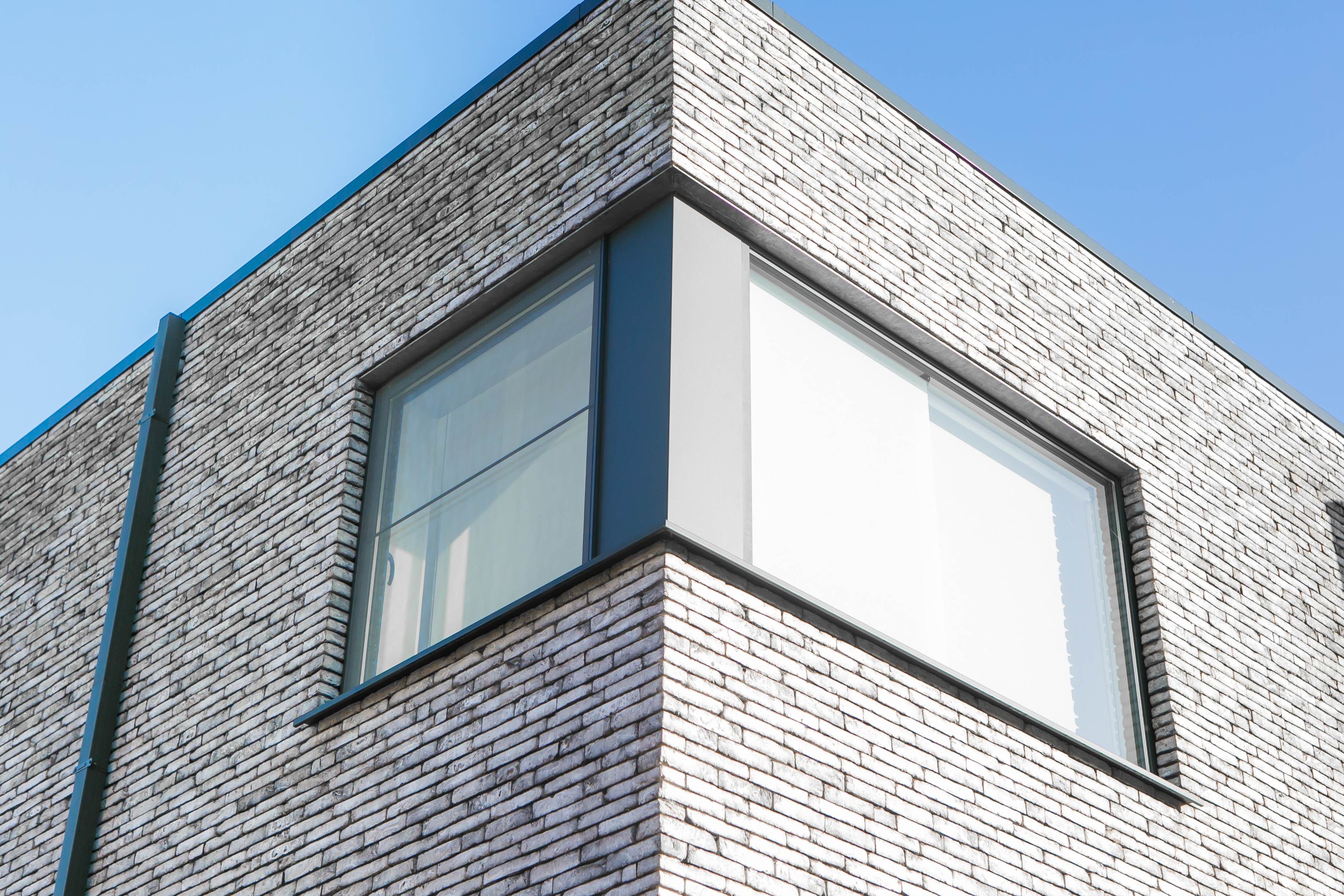 Modern hoekraam met vast en draai-kipgedeelte in zwartgrijze raamprofielen, geïntegreerd in de lichte bakstenen gevel van een nieuwbouwwoning in open bebouwing.