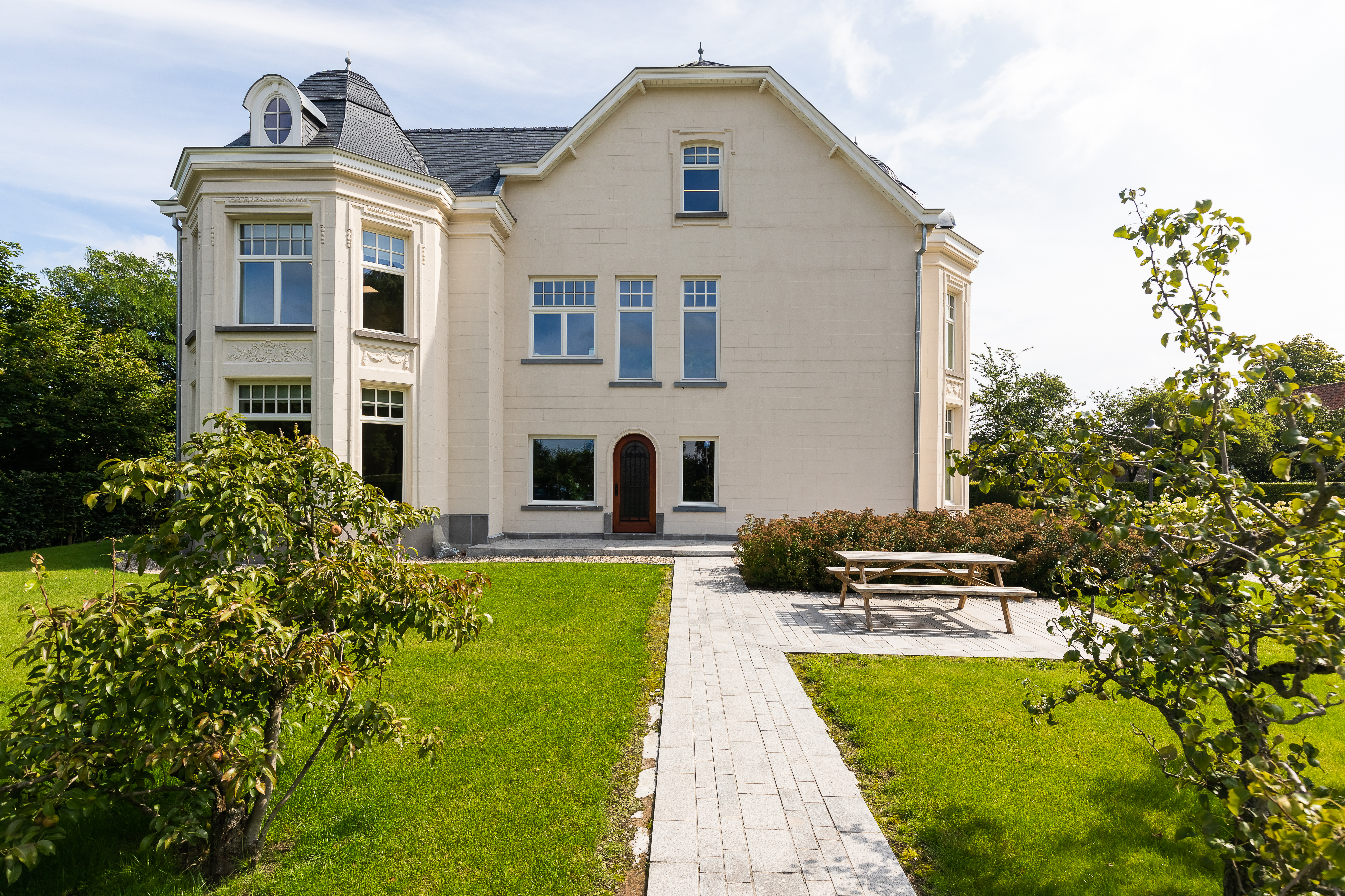 Gerenoveerde open bebouwing in verfijnde klassieke stijl, met witte gevel, hoge ramen, tuin, verhard tuinpad en zitbank.
