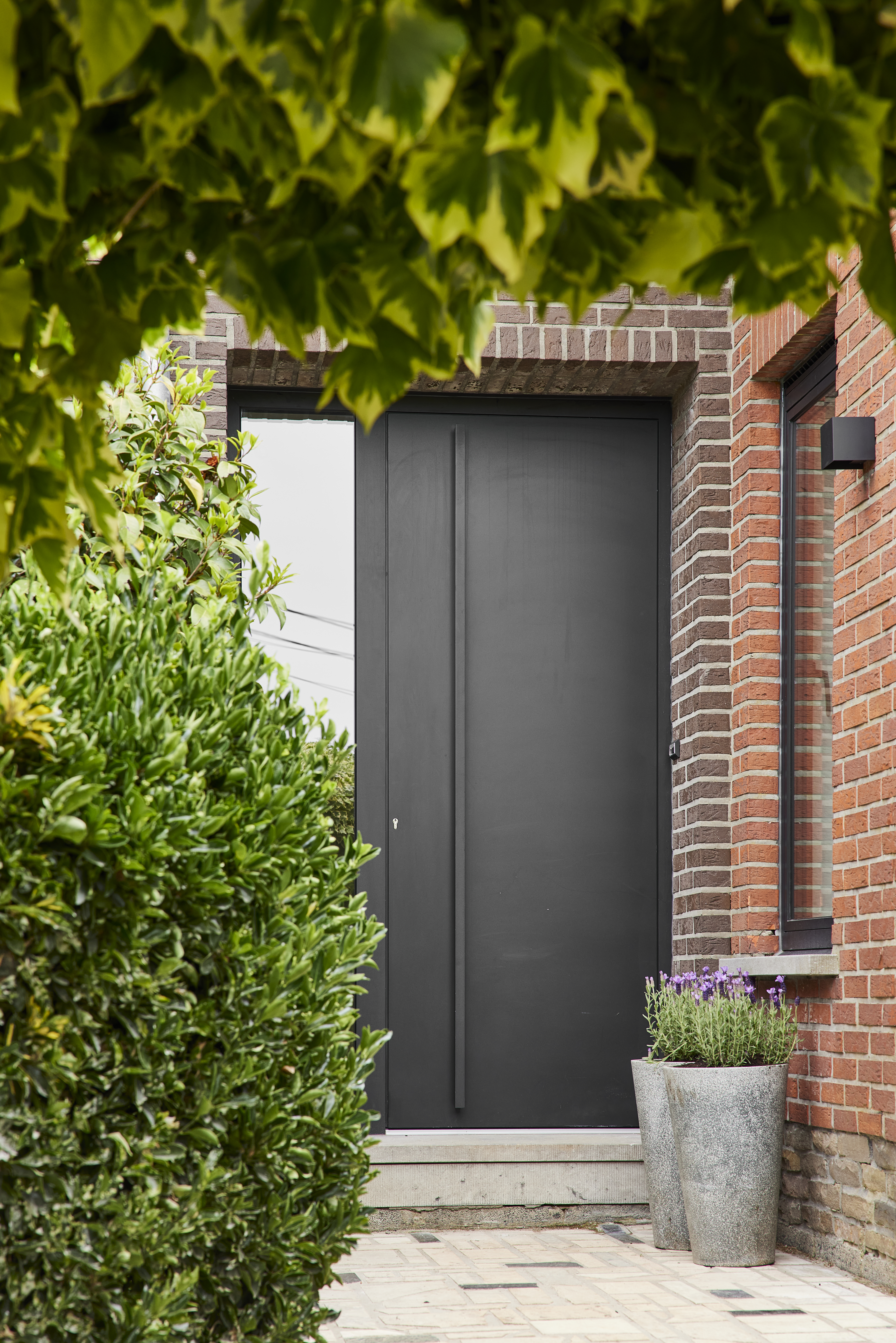 Grafietzwarte aluminiumdeur met zijlicht en paneel in een rode bakstenen gevel, omgeven door groen en een betegeld tuinpad.