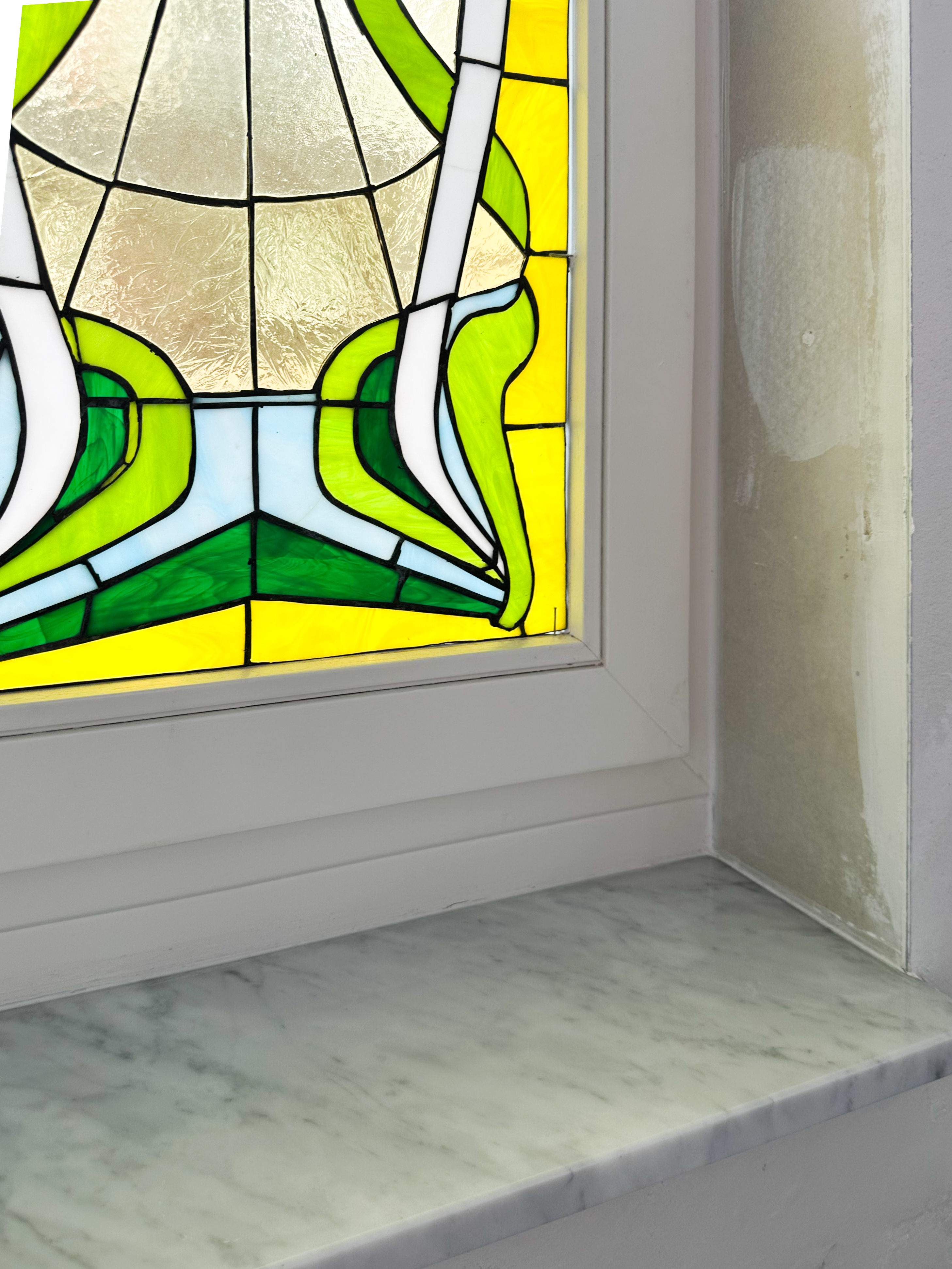 Detail van een vast raam met klassiek glas-in-lood in een wit afgelakt raamprofiel, naast een stenen vensterbank en een bepleisterde muur.