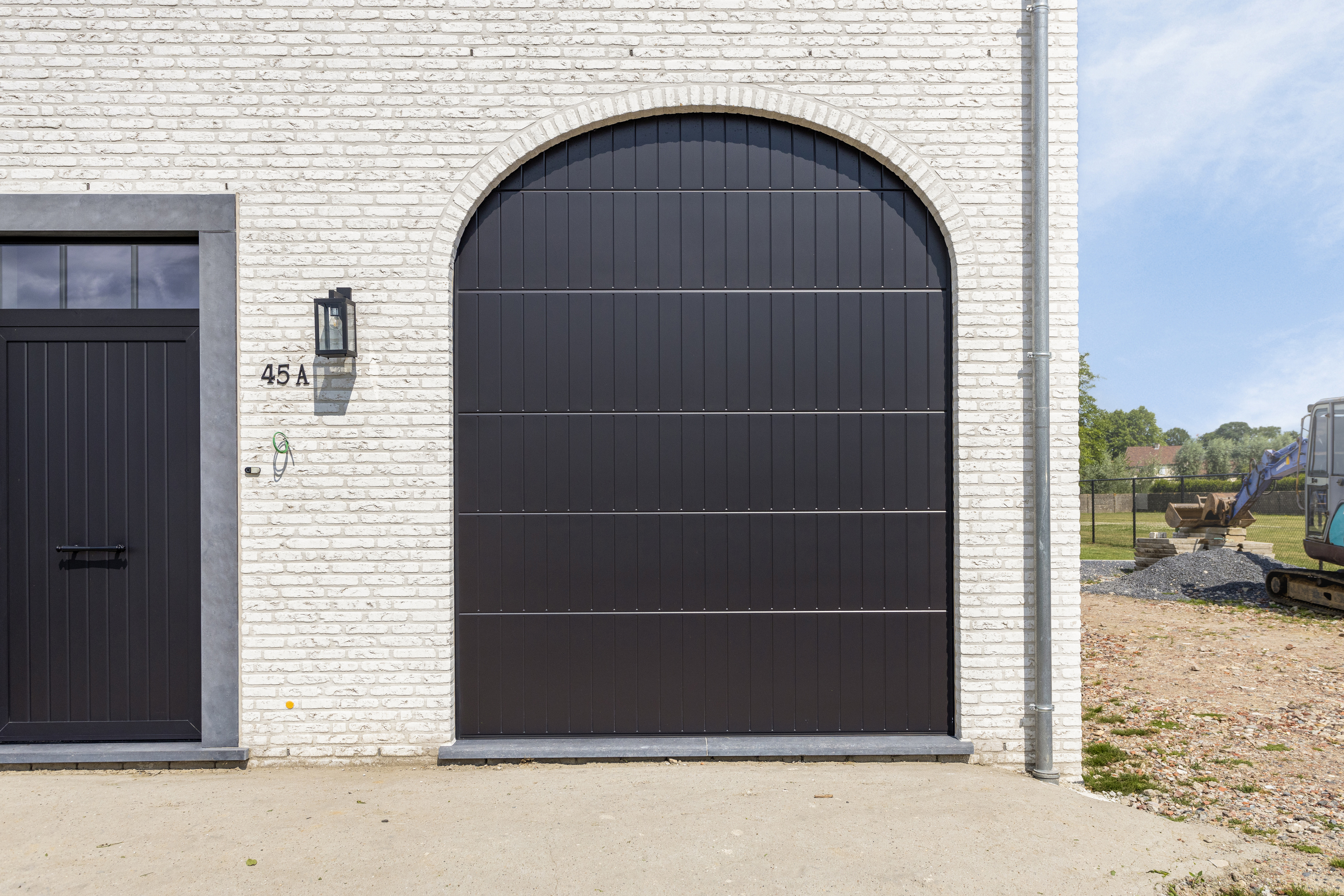 Zwart gebogen garagedeur en bijpassende zijdeur in een lichte bakstenen gevel van een landelijke nieuwbouwwoning.
