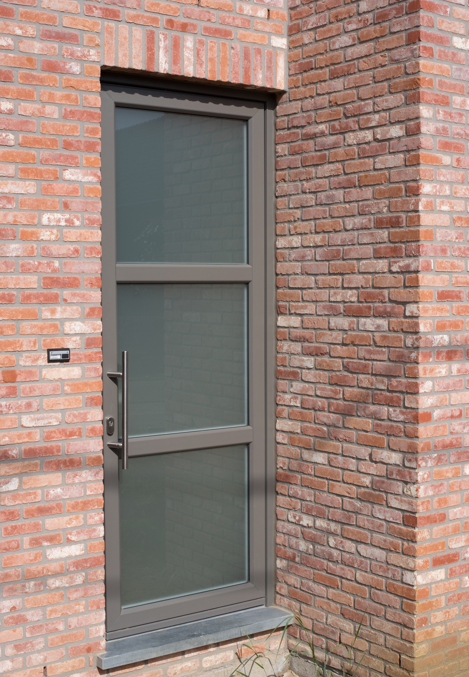 Beglaasde deur in kwartsgrijs met tussenstijl en Sablé-afwerking, geplaatst in een rode bakstenen gevel van een tijdloze nieuwbouwwoning.