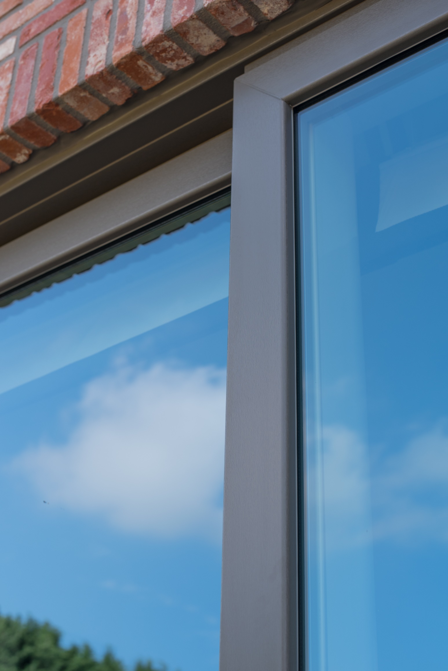 Close-up van een raam met kwartsgrijze NERF-raamprofielen in een bakstenen gevel, met een beglazing die de blauwe lucht en wolken weerspiegelt.