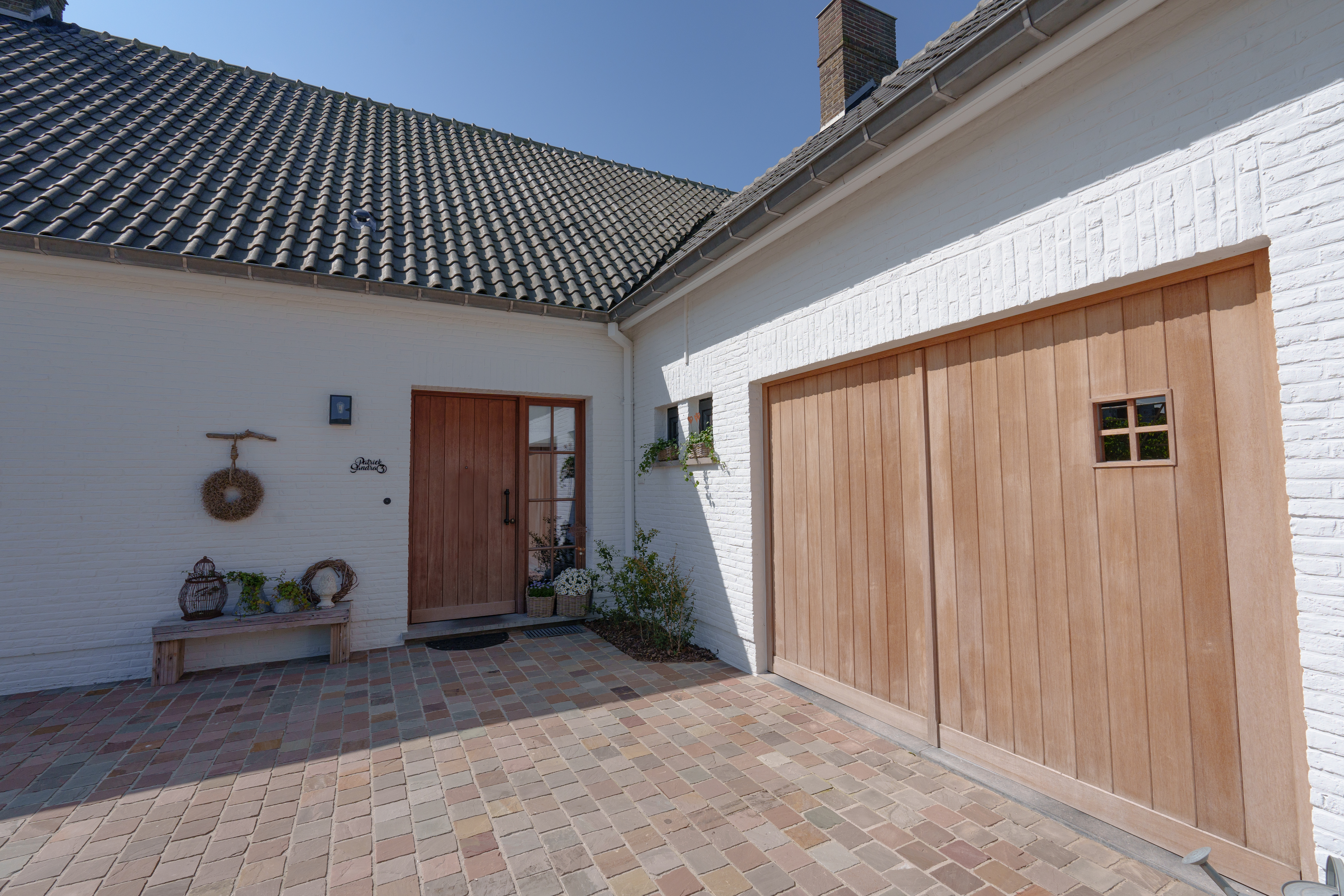 Witte binnenkoer met houten voordeur en zijlicht naast een grote houten garagepoort, onder een pannendak.